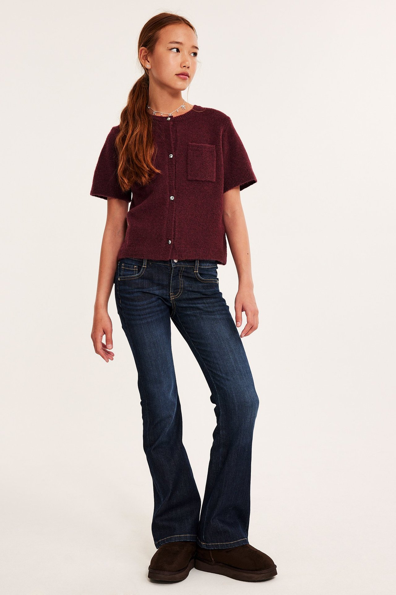 Framifrån: Barn bär en melerad vinröd kortärmad kofta med ficka, mörkblå utsvängda jeans och bruna skor.