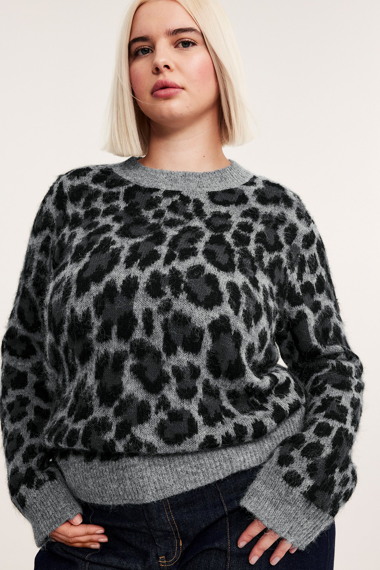 Lähikuva edestä naisesta, jolla on harmaameleerattu jacquard-neulottu leopardikuvioinen neule ja tummansiniset farkut.