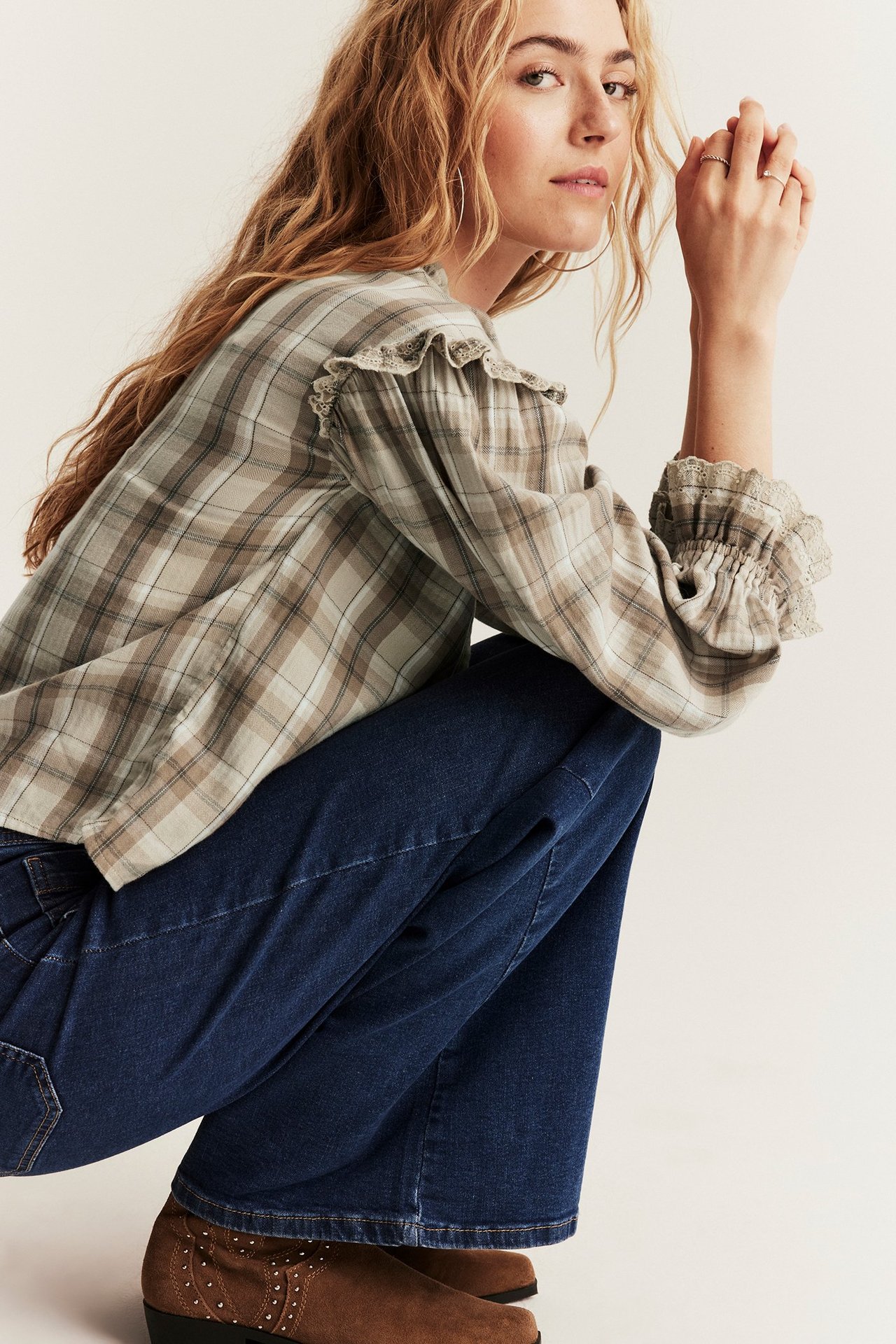 Vinklad framifrån bild av kvinna i beige rutig blus med volanger, mörkblå jeans och bruna stövlar.