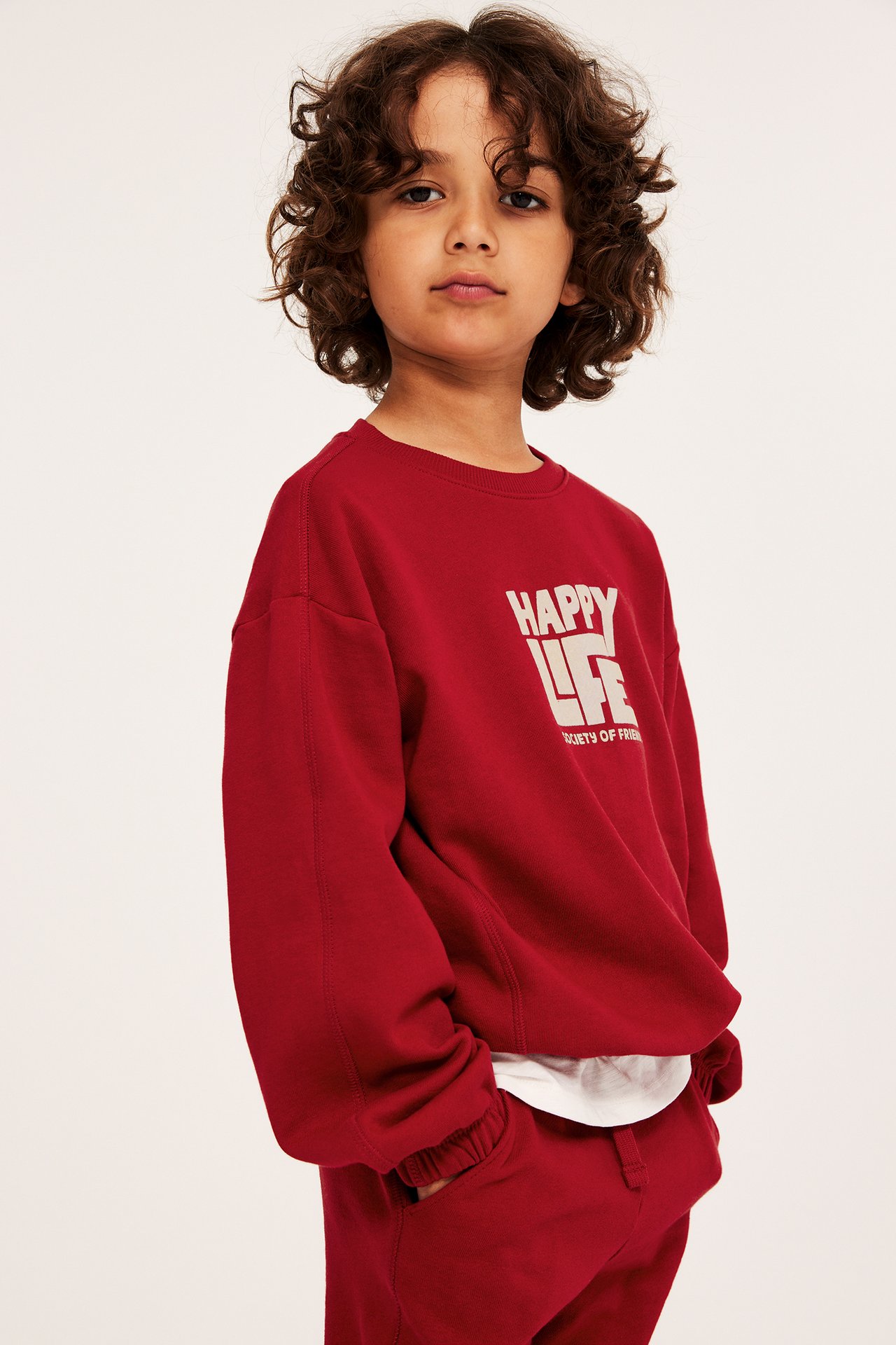 Framifrån, ett barn i röd bomullssweatshirt med "HAPPY LIFE"-tryck, över röda mjukisbyxor.