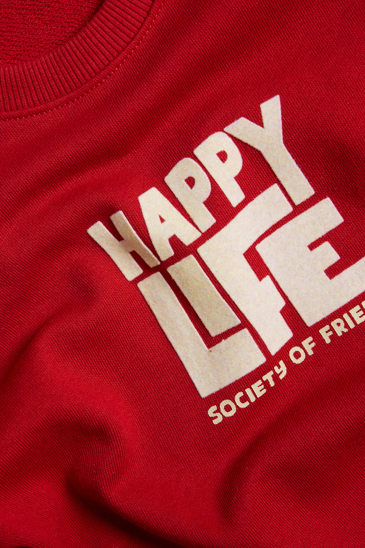 Närbild på en röd bomullssweatshirt med vitt velourtryck "HAPPY LIFE" framtill.