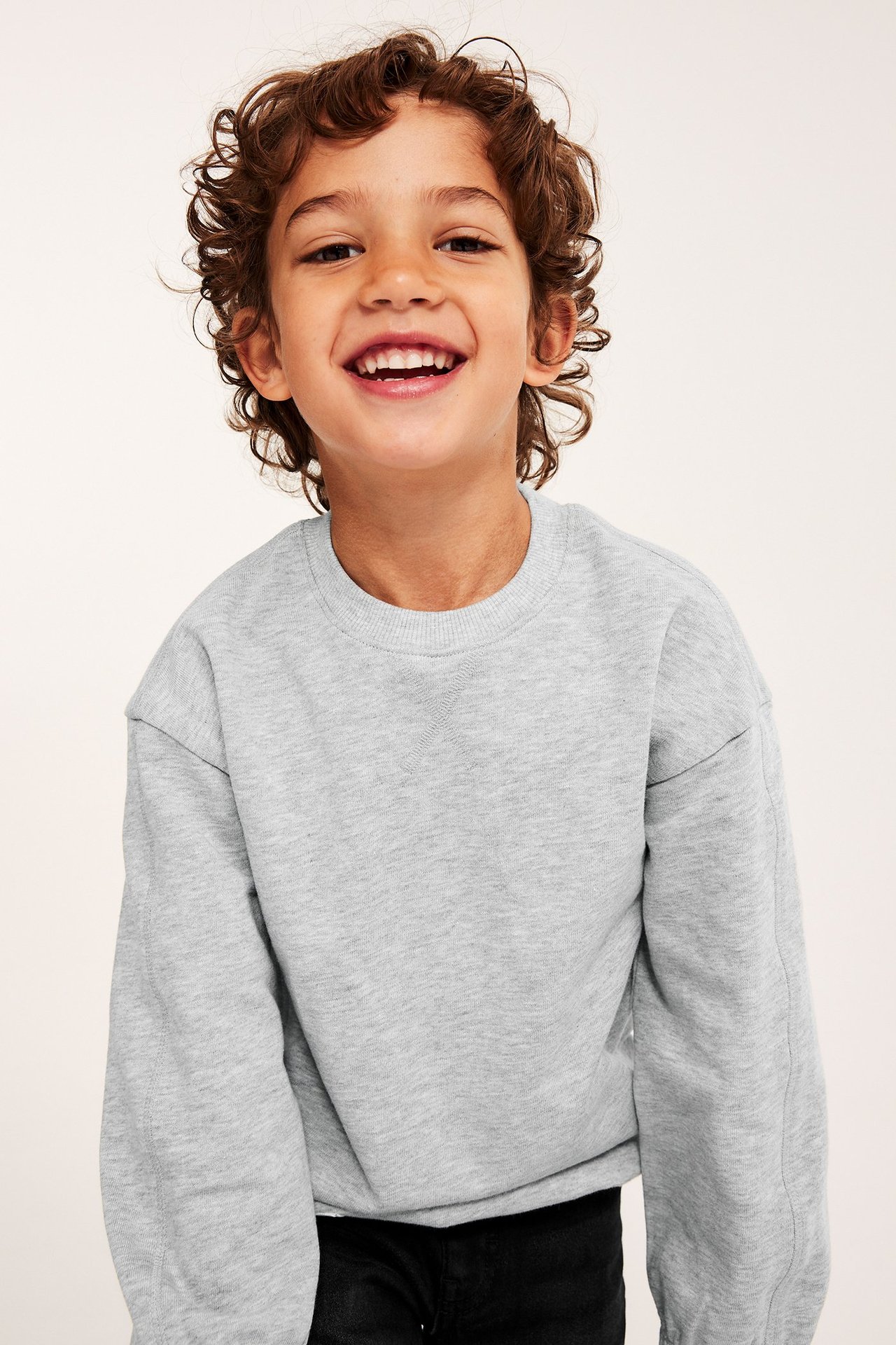 Framifrån, ett barn i en ljusgrå bomullssweatshirt med ribbad halsringning, bär svarta byxor.