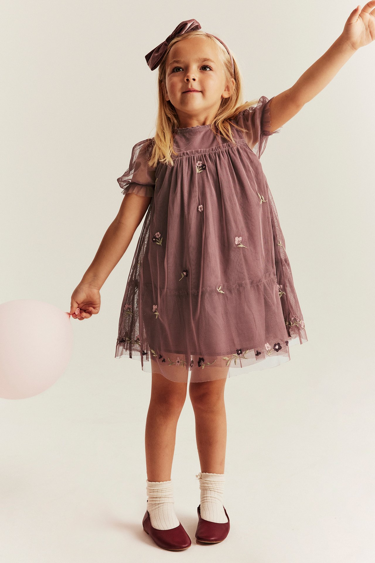 Frontvy: Kid i mörkrosa broderad nätklänning med puffärm, sammetspannband, vita strumpor och håller en ballong.