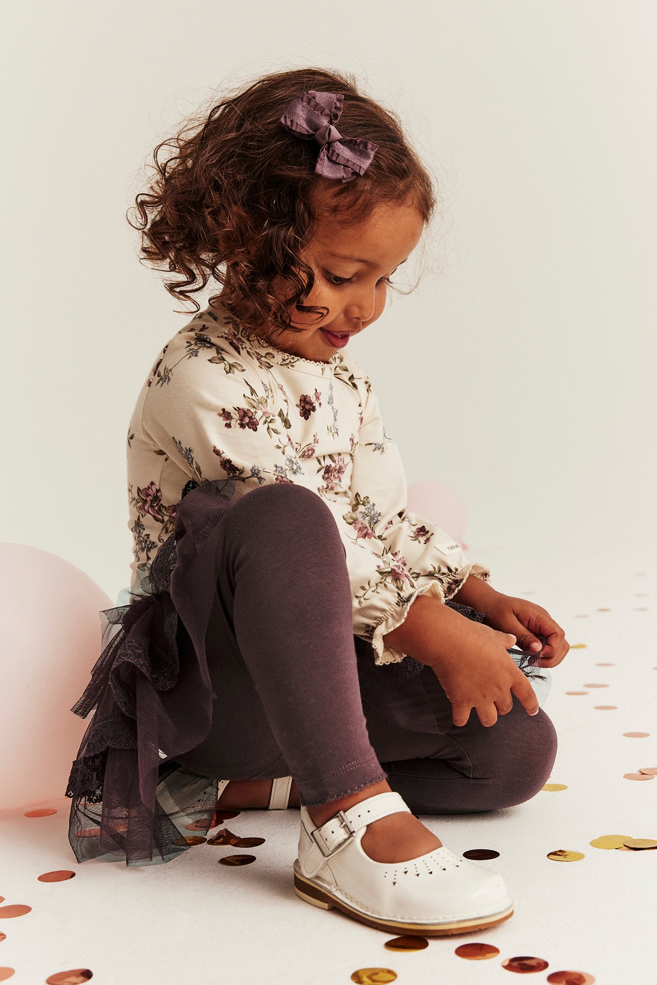 Sidovy: Barn bär syrenlila leggings, volangkjol i tyll, blommig topp, vita skor, lila rosett.