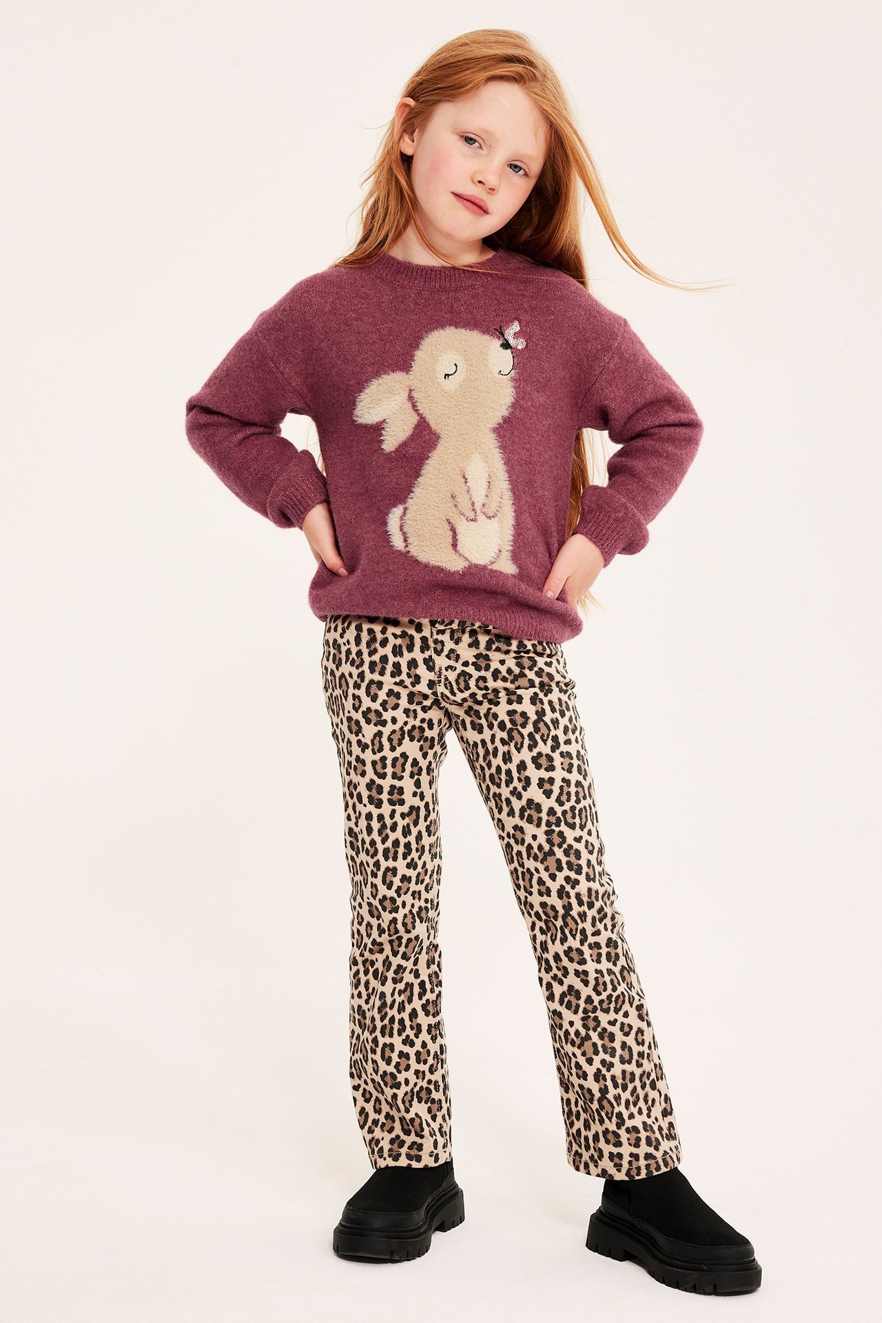 Widok z przodu: Dziecko w ciemnoróżowym swetrze z królikiem, spodniach w panterkę i czarnych masywnych butach.