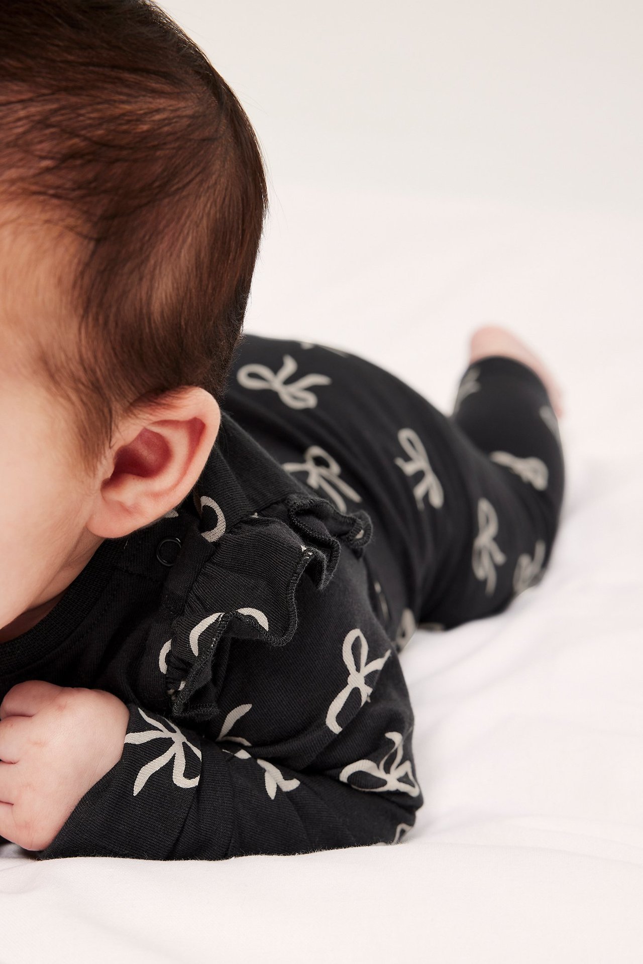 Närbild sidovy av baby i svart mönstrad bomullsbody med vita rosetter och axelvolanger.