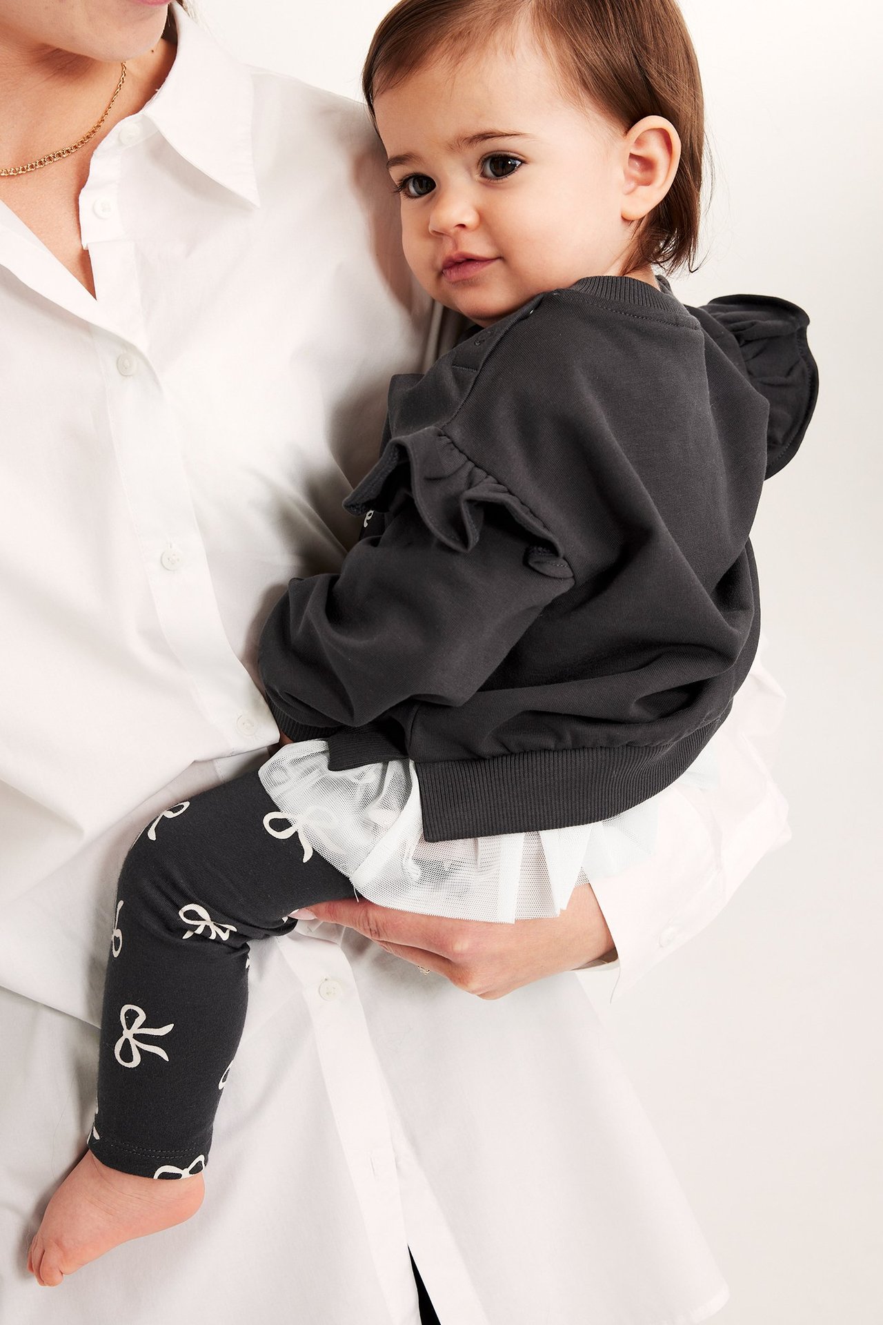 Närbild på en baby i svart sweatshirt med axelvolanger, tryck fram, vit meshfåll och svarta leggings med rosettmönster.