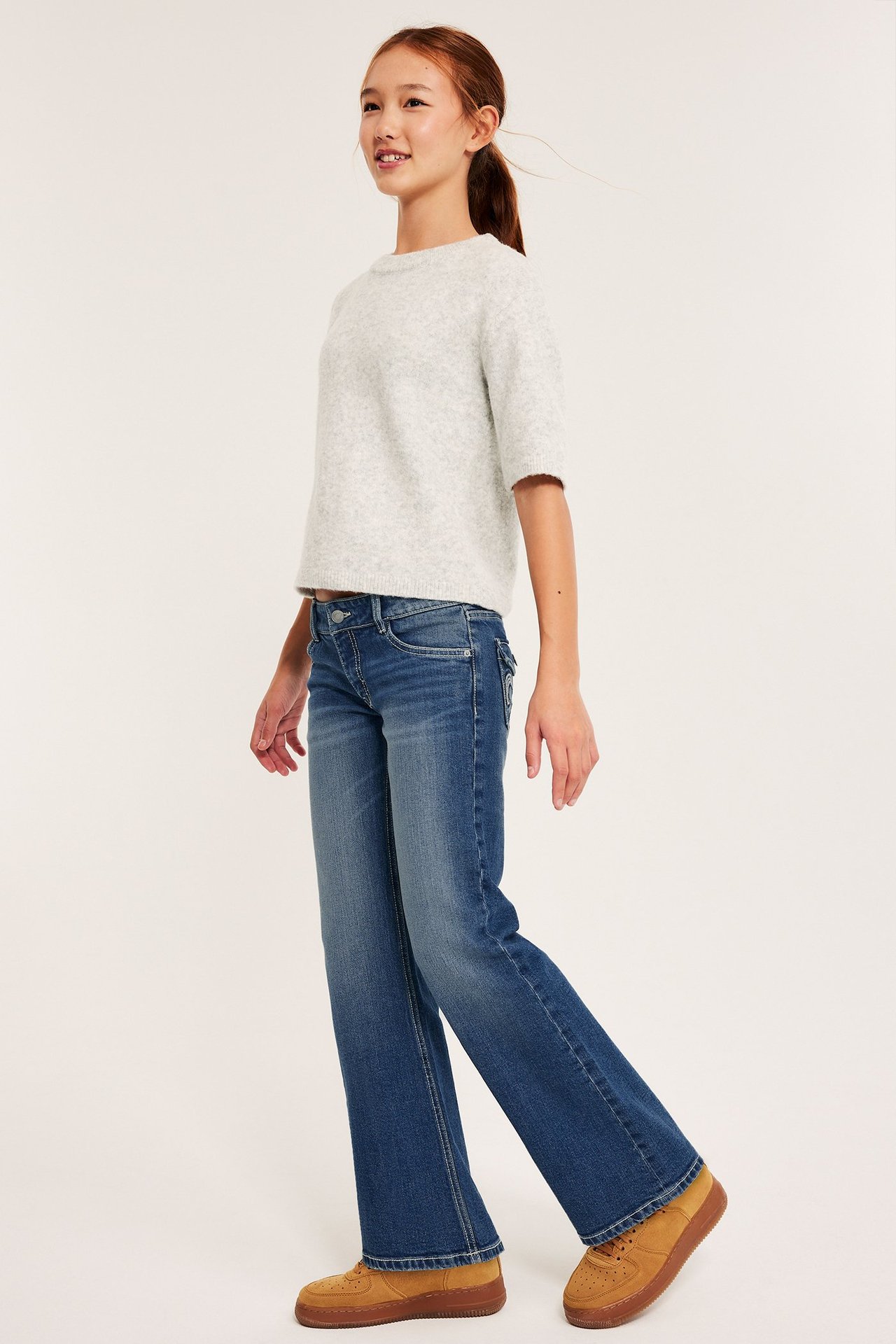 Flicka i vit tröja och blå bootcut jeans med broderade bakfickor, framifrån.