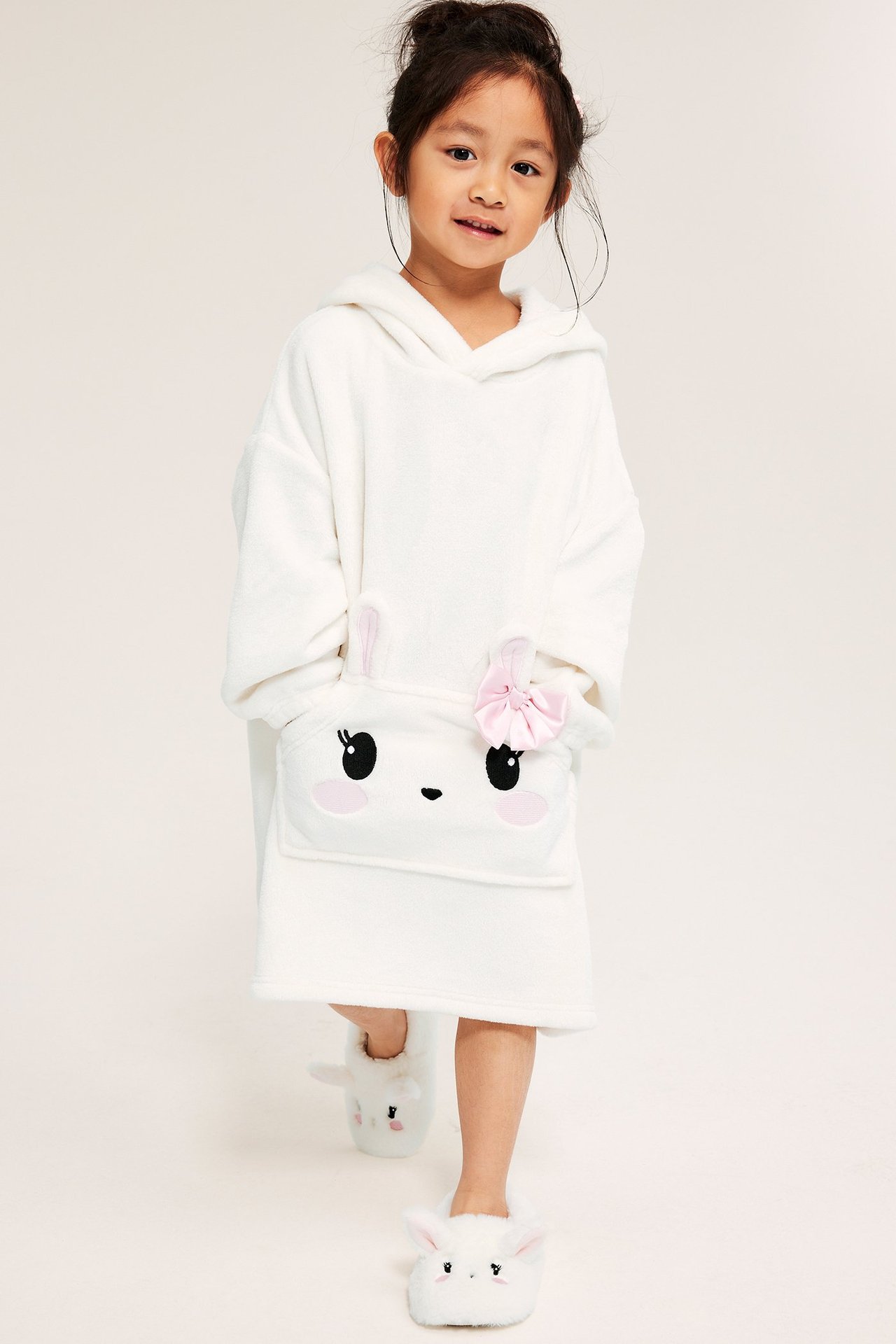 Dziewczynka w białej polarowej bluzie z króliczym pyskiem, różową kokardką, z przodu.