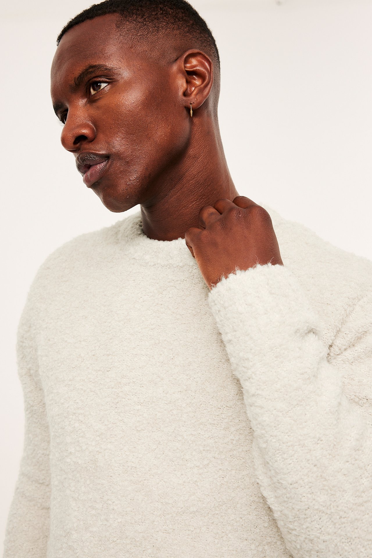 Sideprofil, mann i en beige bouclé O-hals genser, med en liten gullfarget bøyleørering.