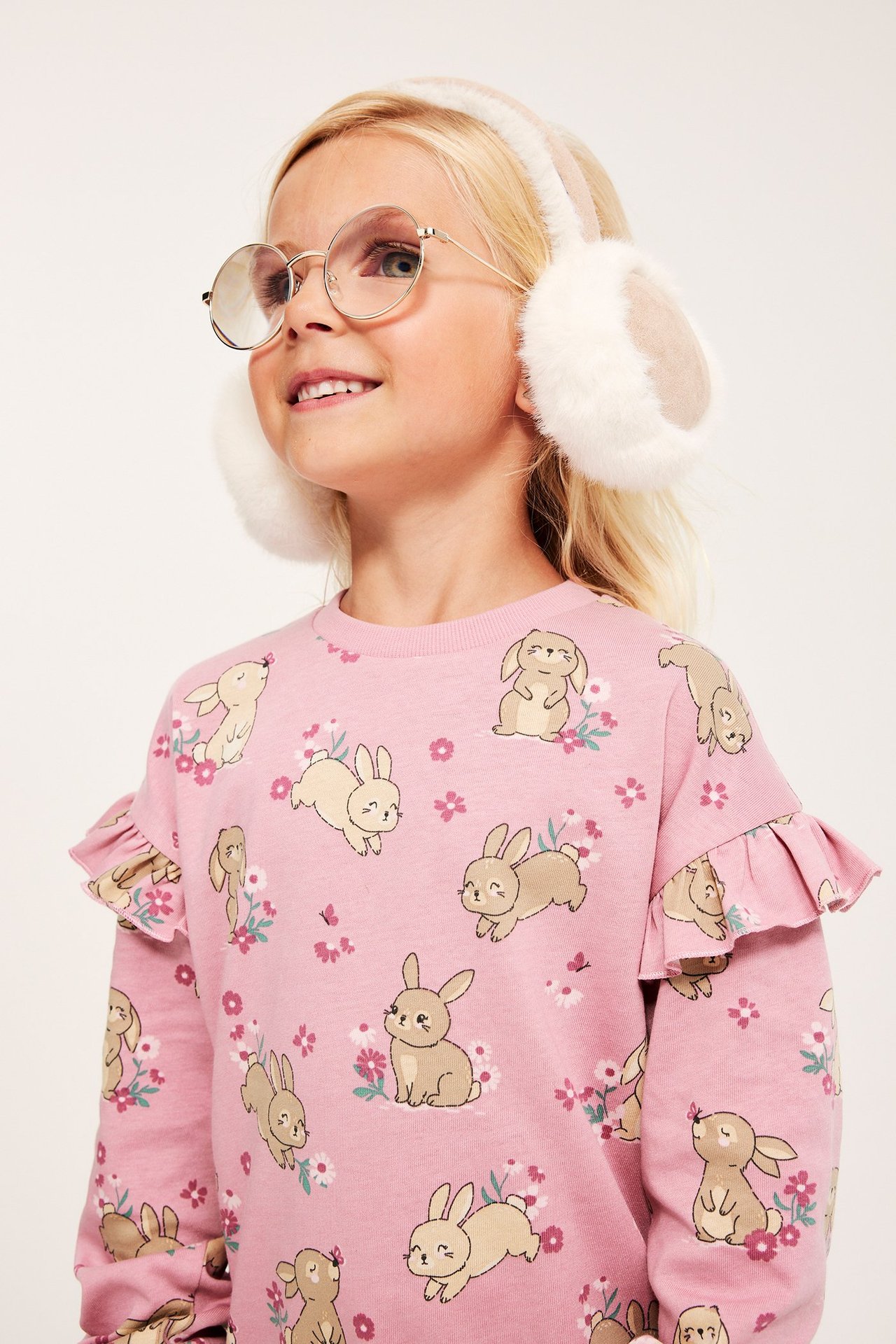 Nærbilde av et barn i rosa bomullssweatshirt med kanintrykk, volangermer, runde briller og hvite øreklokker.