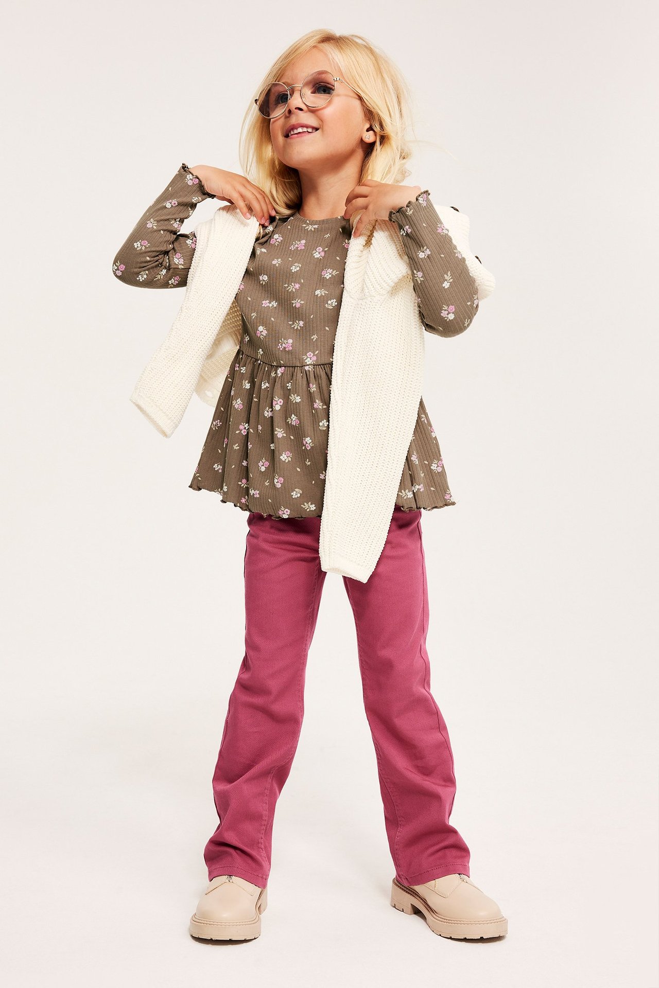 Frontbilde av et barn i en beige ribbet blomstret peplumtopp, hvit cardigan, rosa bukser og beige sko.