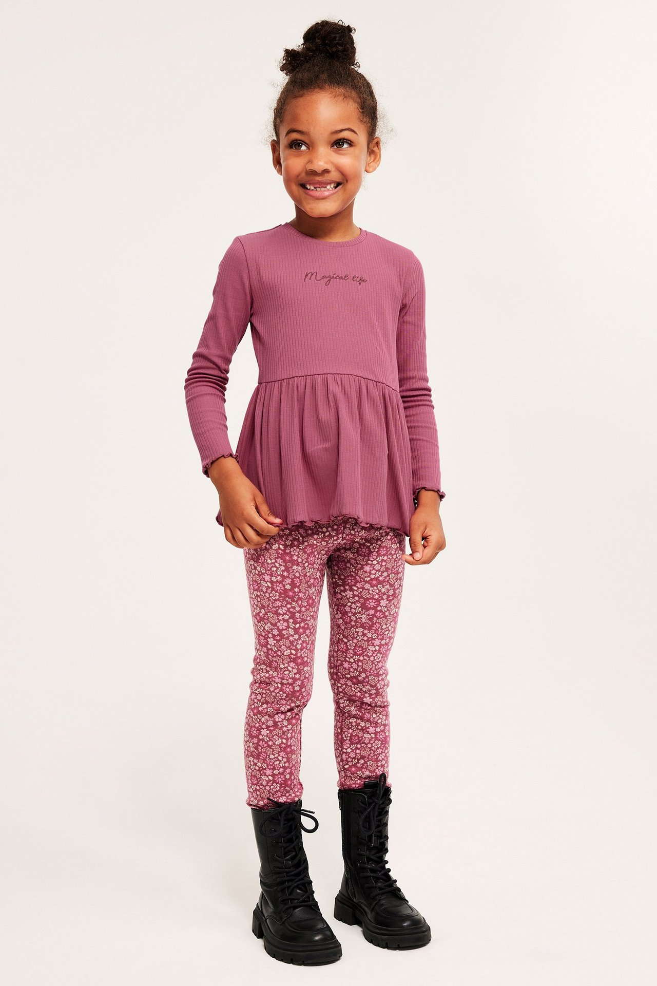 Edestäpäin lapsi yllään tummanpunainen ribattu peplumtoppi tekstillä, kukkakuosiset leggingsit ja mustat saappaat.