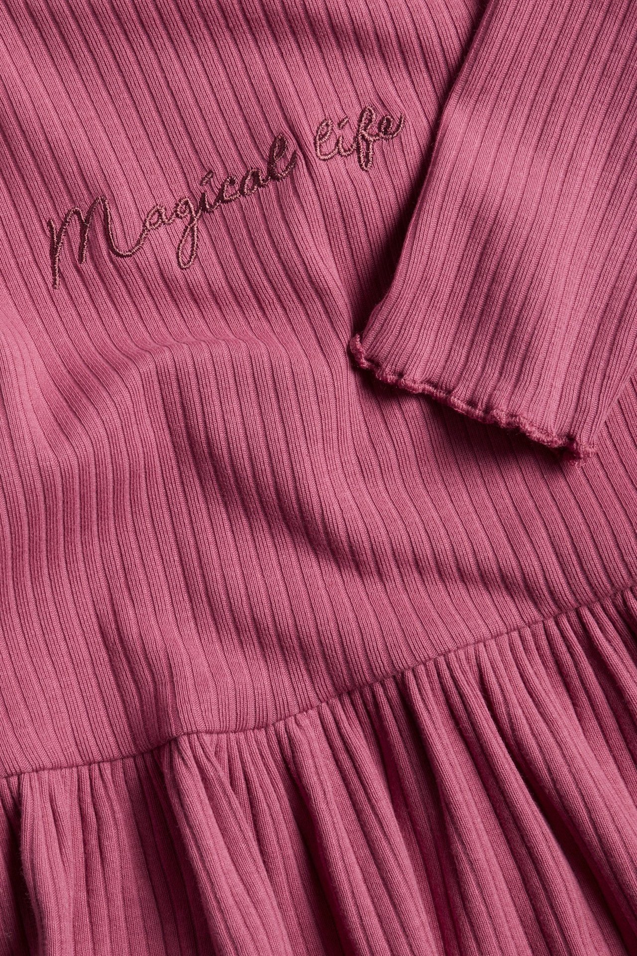 Zbliżenie na ciemnoróżowy prążkowany top peplum z haftem "Magical life" i falbankowymi mankietami.