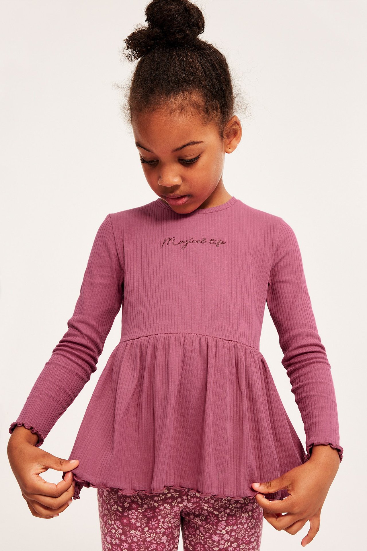Widok z przodu dziecka w ciemnoróżowym prążkowanym topie peplum z haftem "Magical life" i kwiecistych legginsach.