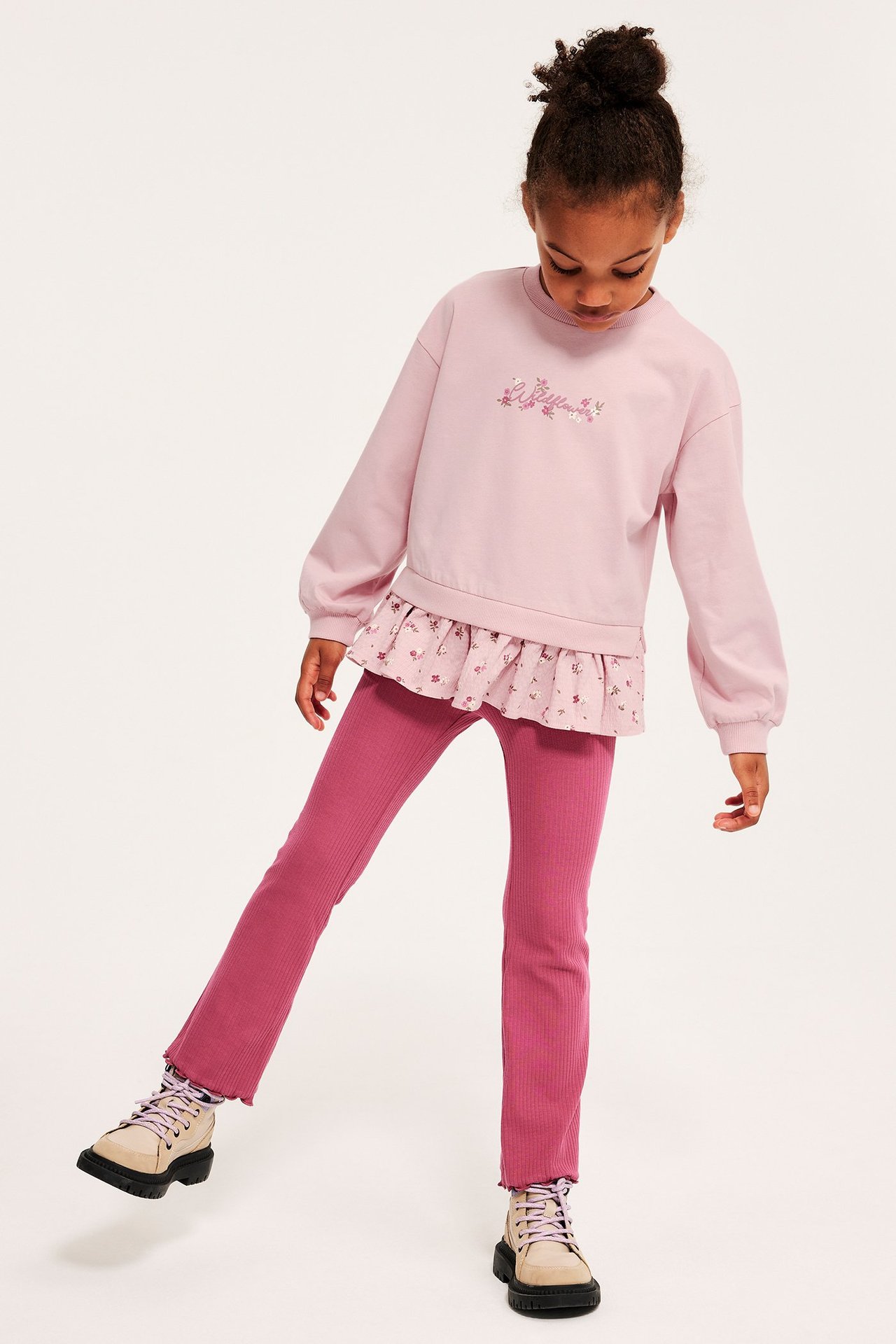 Frontbilde av et barn i rosa sweatshirt med blomsterbroderi, blomstret peplum, magentaribbet leggings og beige støvler.