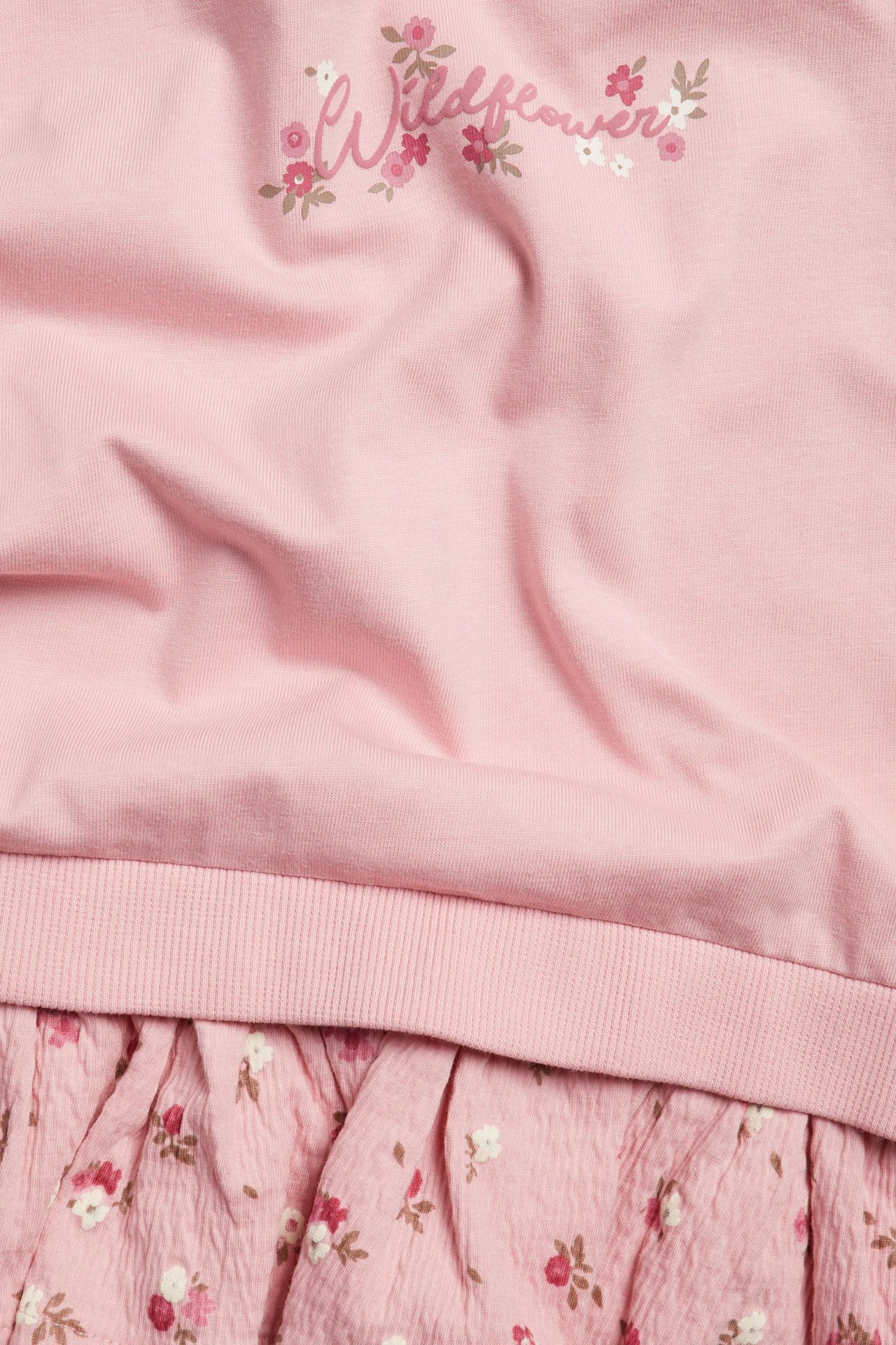 Zbliżenie na jasnoróżową bluzę dziecięcą z baskinką, z nadrukiem "Wildflower" i kwiecistą falbanką.