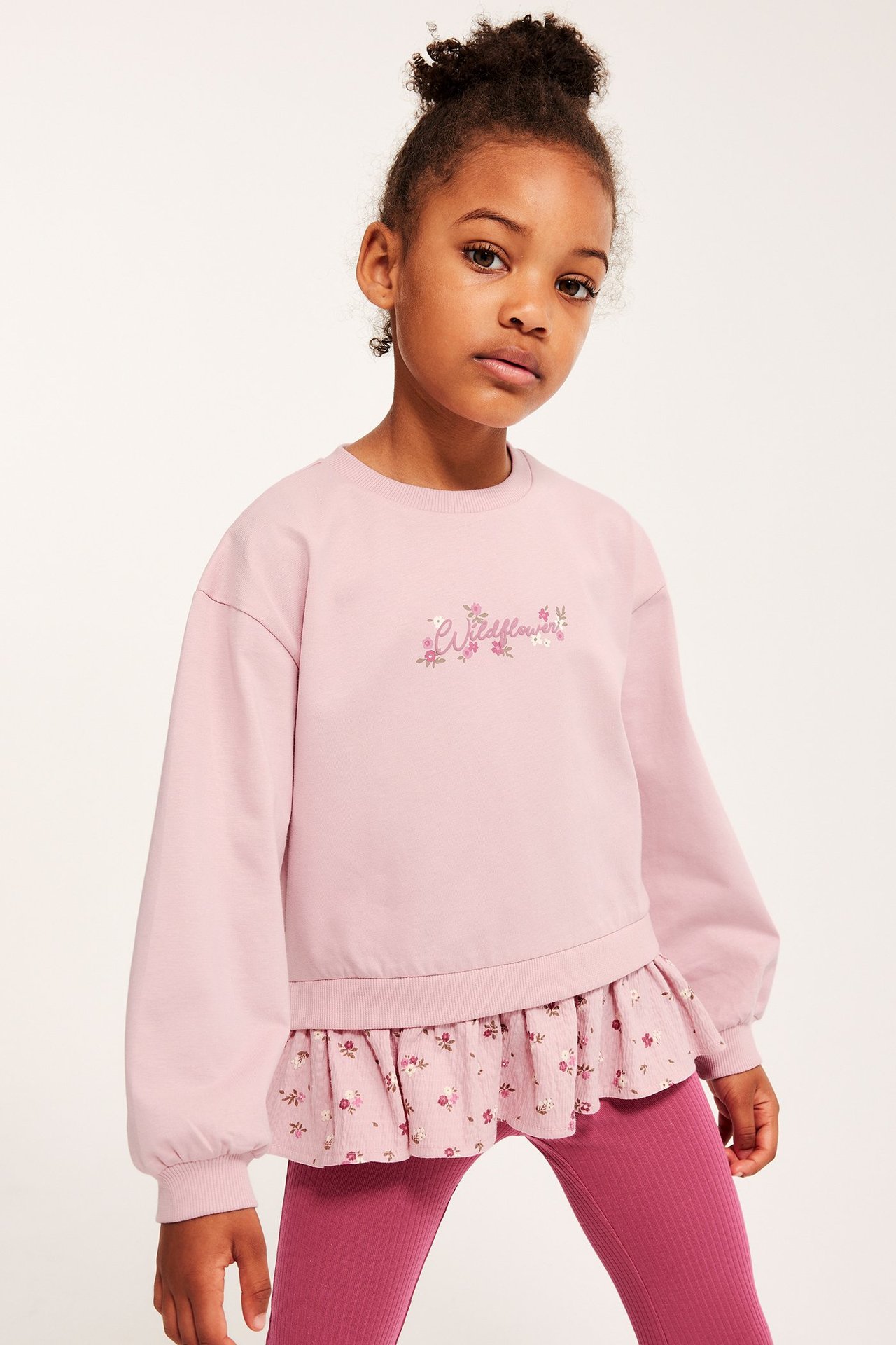 Widok z przodu dziecka w jasnoróżowej bluzie z kwiatowym peplumem i ciemnoróżowych legginsach w prążki.