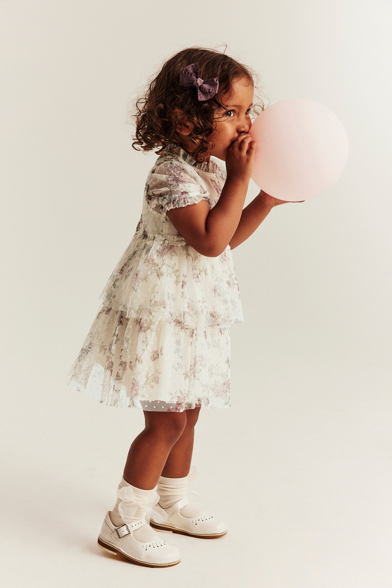 Sidebilde av et barn i lys beige blomstret meshkjole med volanger, blåser en rosa ballong.