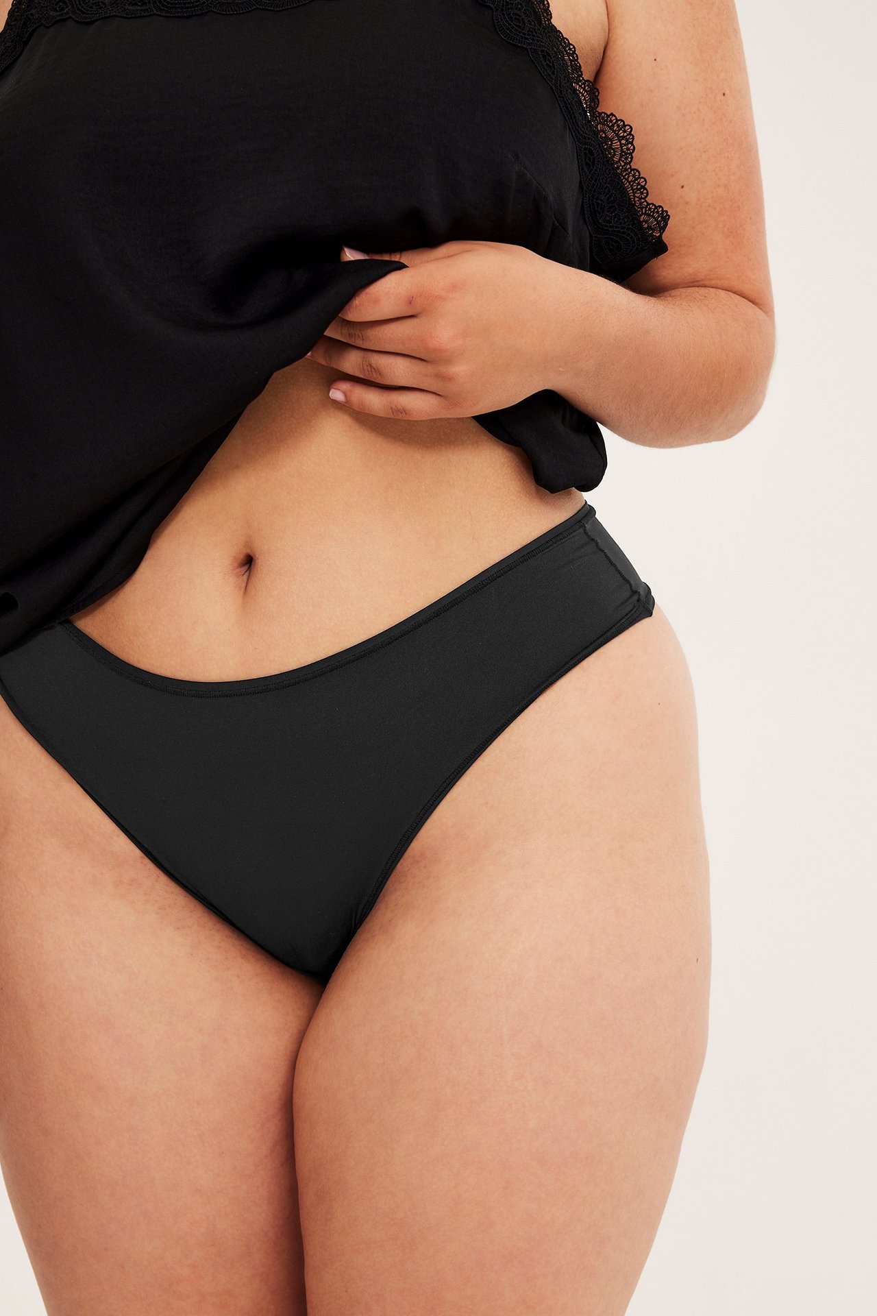 Lähikuva edestä naisesta, jolla on mustat korkeavyötäröiset stringit ja musta pitsikoristeinen toppi.