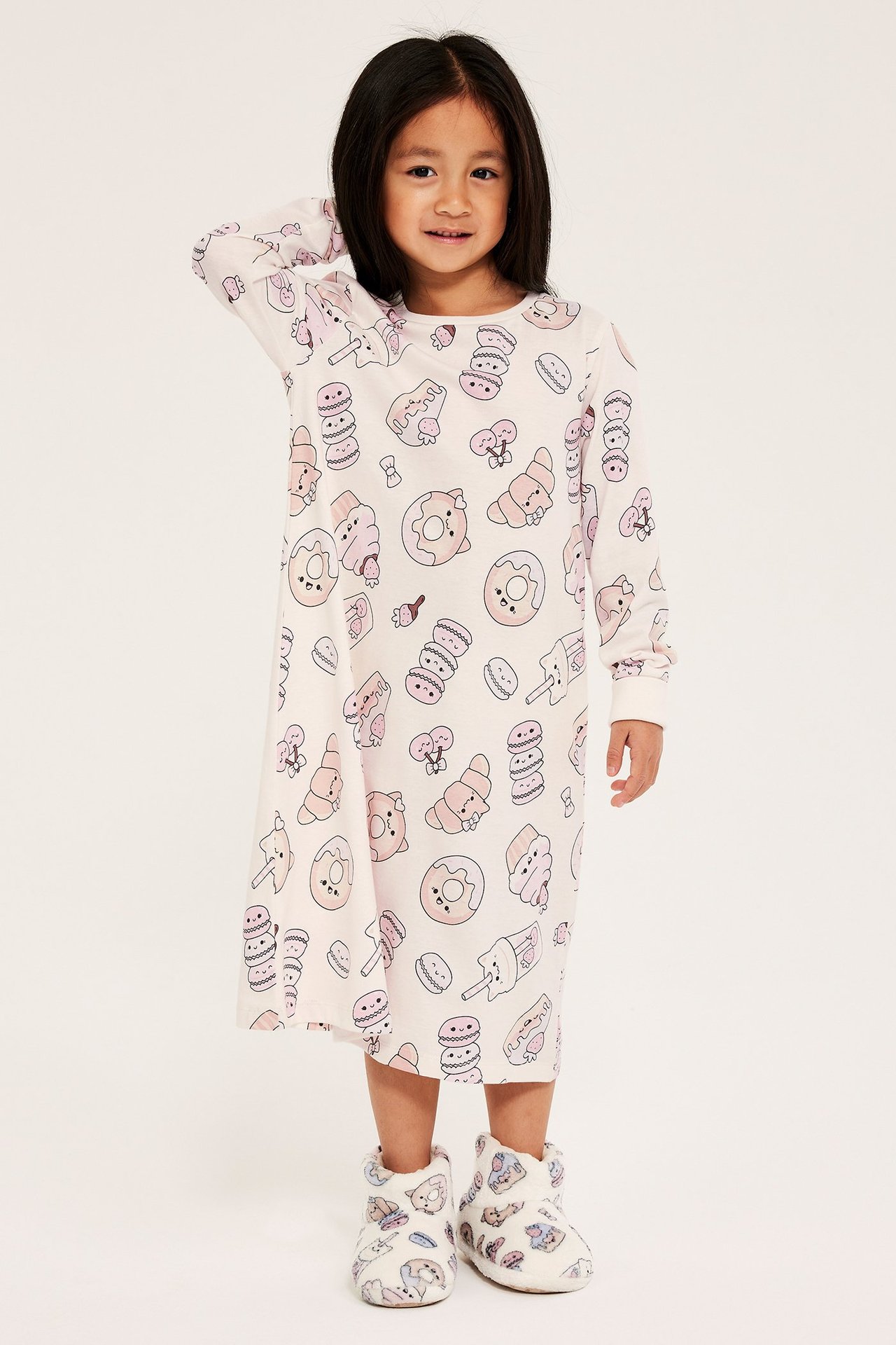 Edestä: Lapsi luonnonvalkoisessa pitkähihaisessa puuvillayöpaidassa jälkiruokakuviolla ja yhteensopivissa tossuissa.