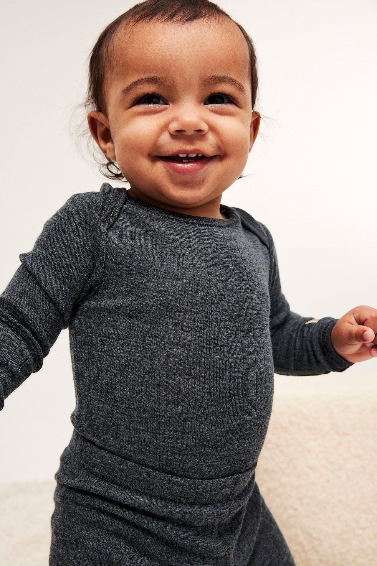 Lähikuva edestä hymyilevästä vauvasta yllään tummanharmaa meleerattu ribattu pitkähihainen villabody olkapään napeilla.