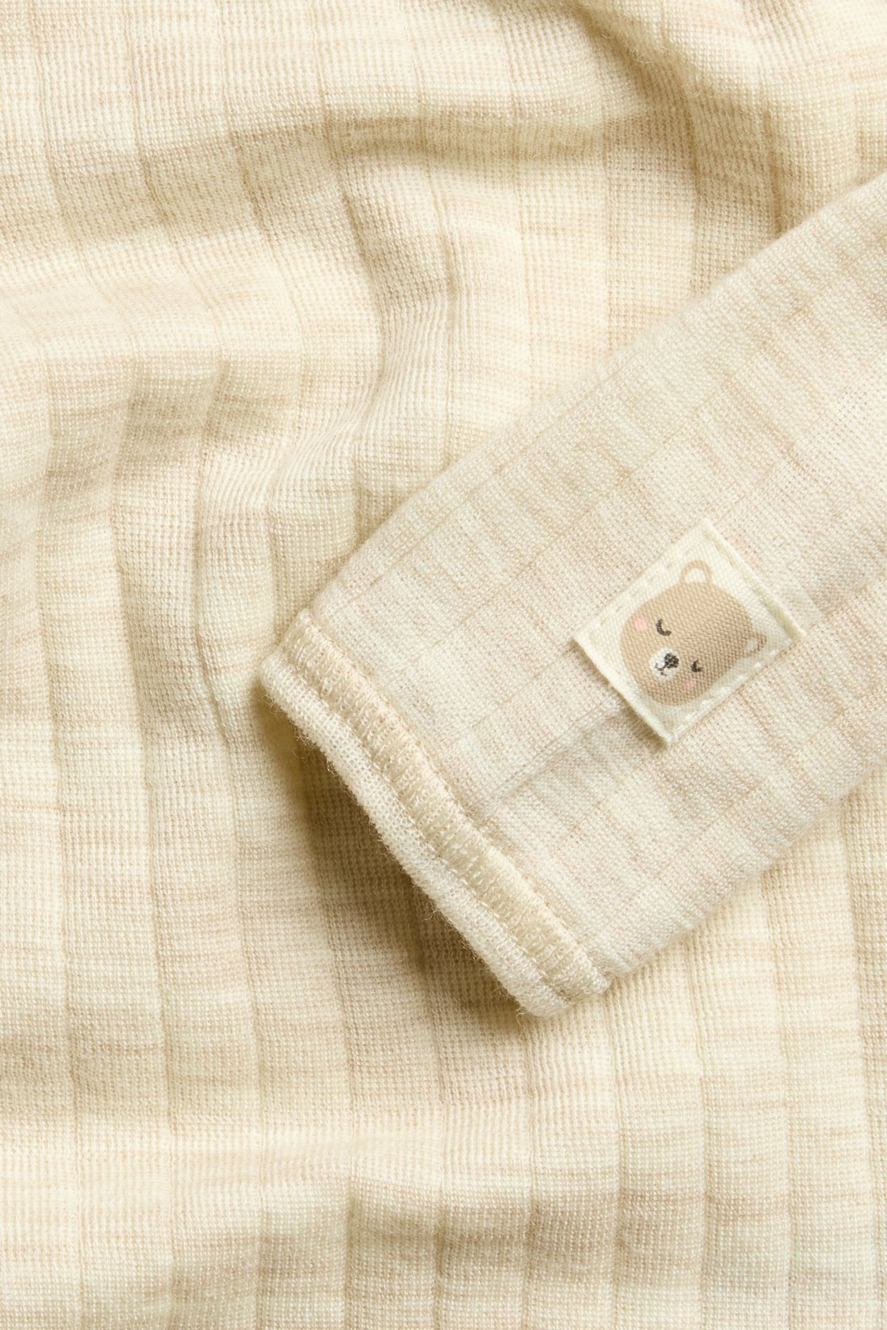 Närbild på en baby beige melerad ribbad ullbody med en sovande björn-patch på ärmen.