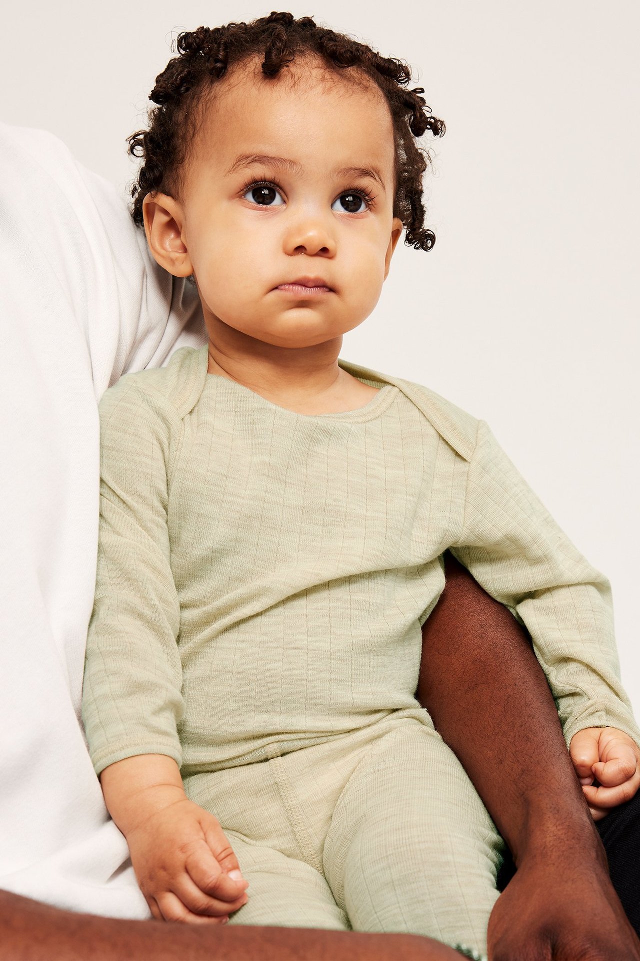 Närbild framifrån på en baby i en ljusbeige melerad ribbad långärmad ullbody och matchande leggings.