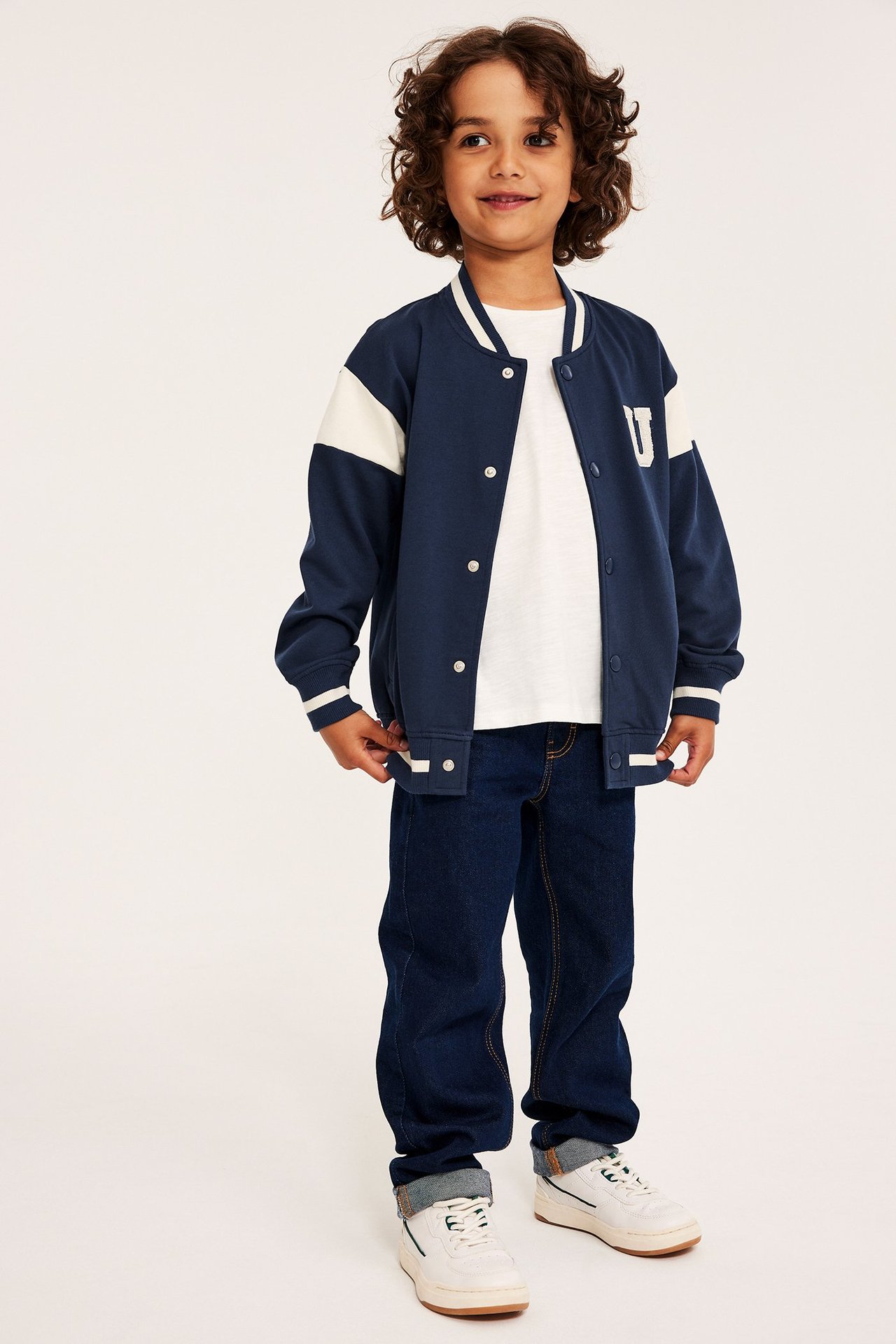 Framifrån: Barn i mörkblå baseballjacka, vita ärmar, 'U'-märke, tryckknappar, vit t-shirt, mörka jeans, sneakers.
