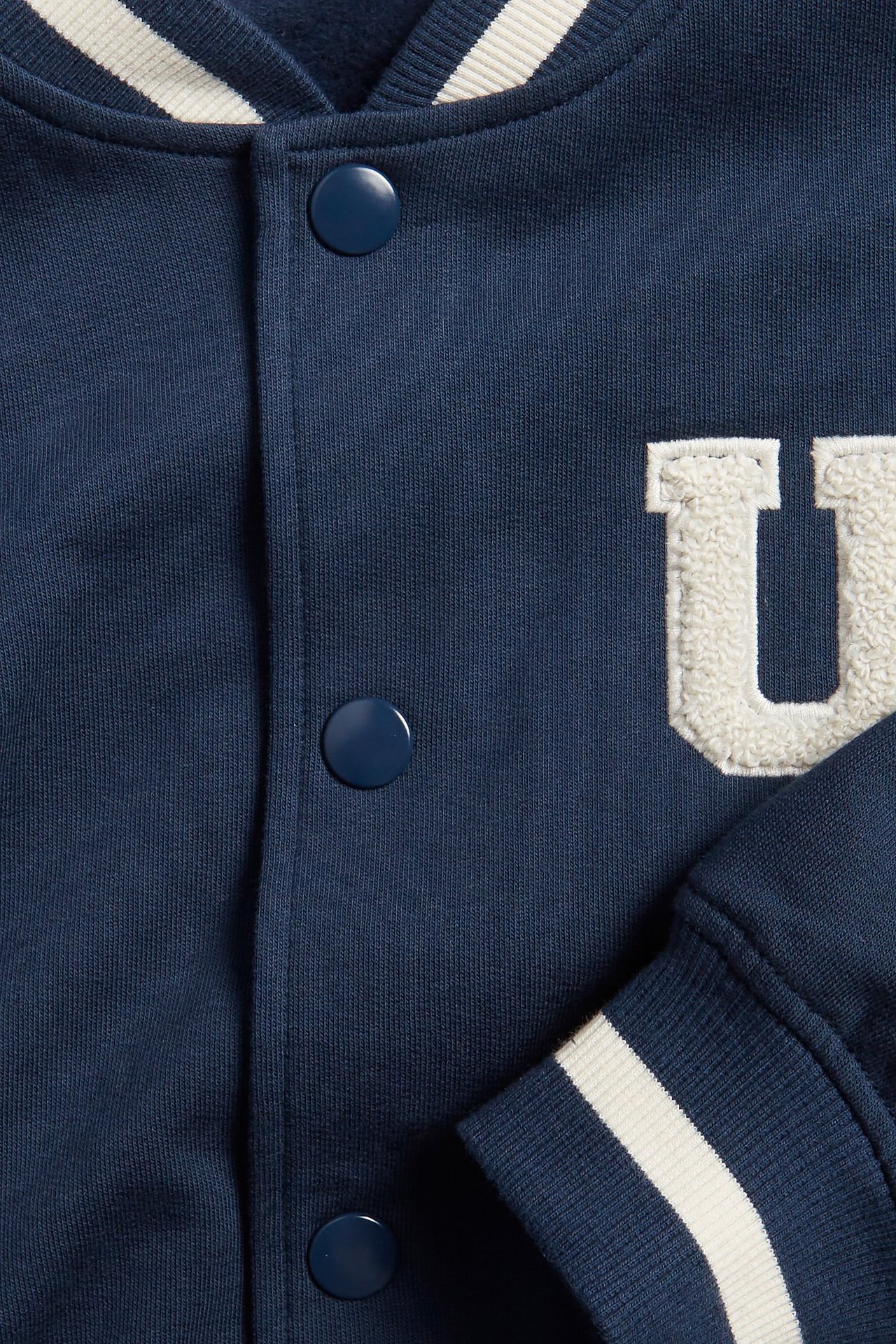 Zbliżenie na granatową bawełnianą kurtkę bejsbolówkę z białymi paskami na kołnierzu, mankietach i kremową aplikacją 'U'.