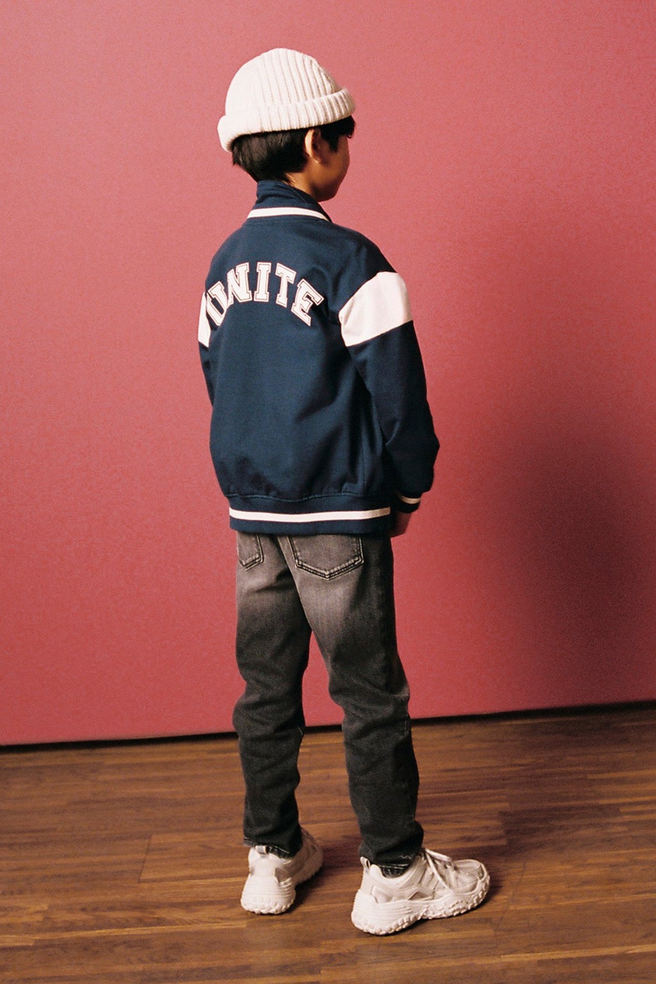 Bakifrån: ett barn i en mörkblå baseballjacka, grå jeans, vit mössa och vita sneakers.