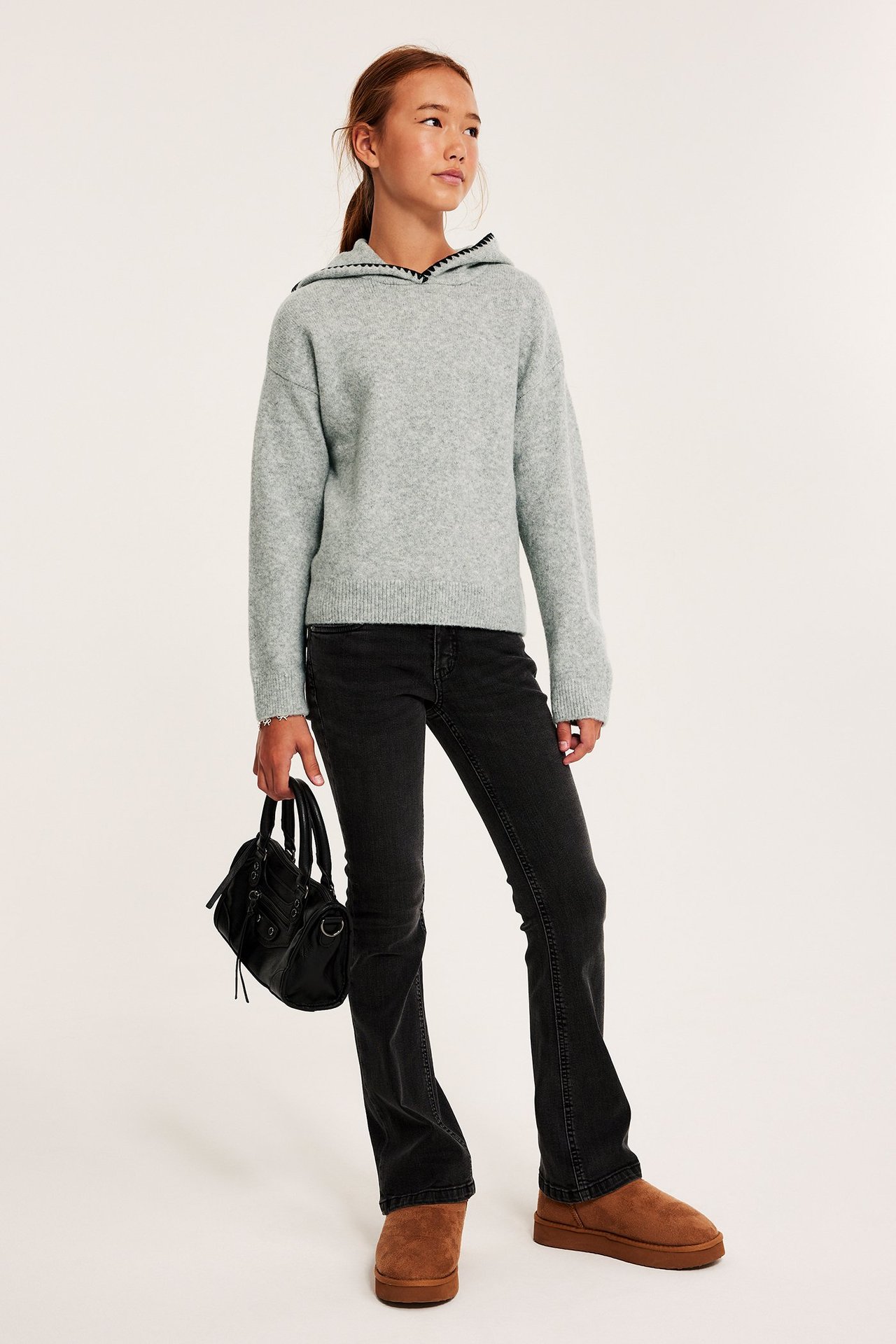 Edestäpäin kuvattu lapsi vaaleanharmaassa melange-neulehupparissa, mustissa farkuissa, ruskeissa saappaissa, pitäen mustaa käsilaukkua.