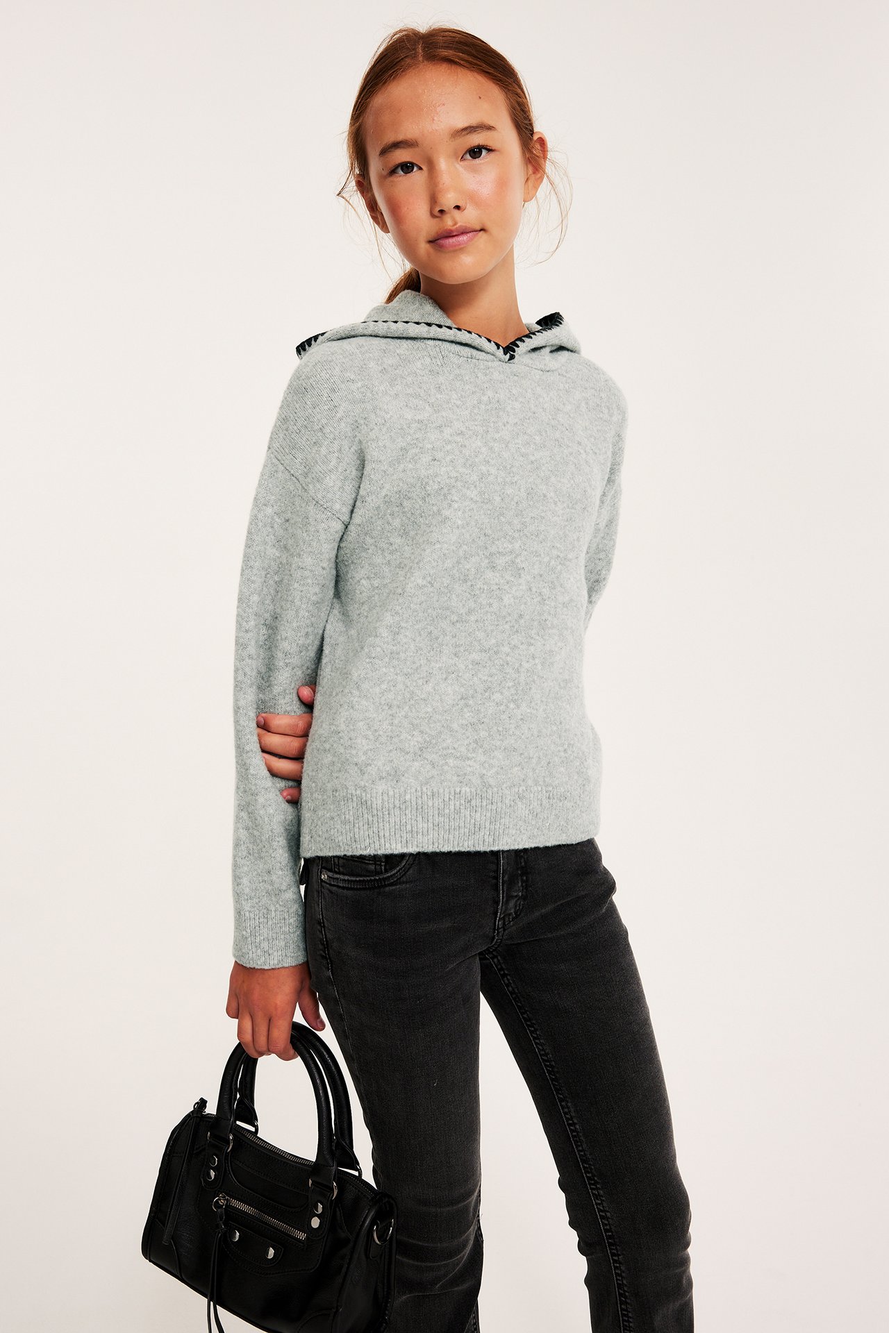 Frontvisning: Barn i lys grå strikket hoodie med dekorative sømmer, svarte jeans, holder en svart håndveske.