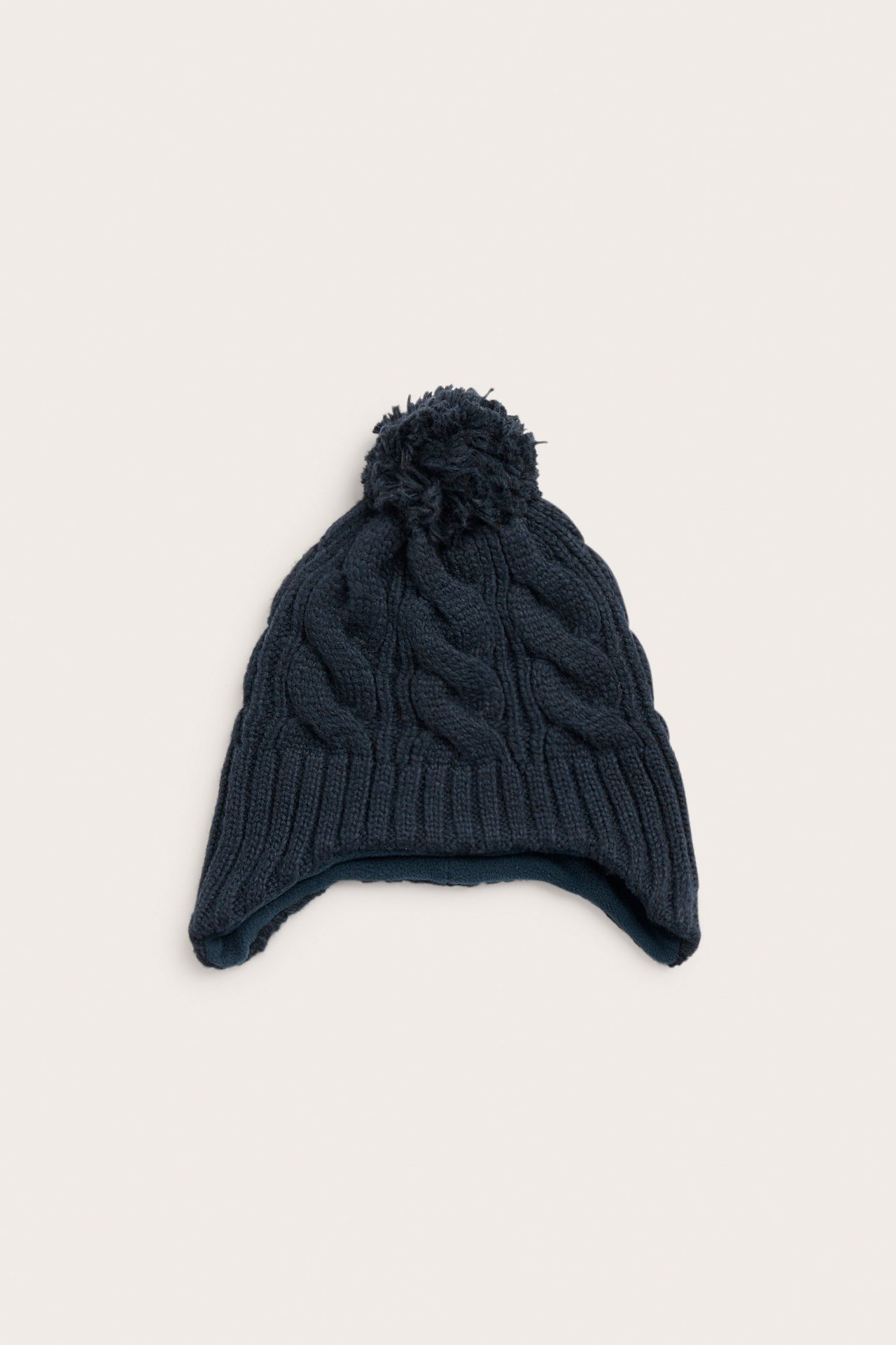 Edestä näkymä tummansinisestä palmikkoneulotusta lasten piposta, jossa on tupsu ja korvaläpät.