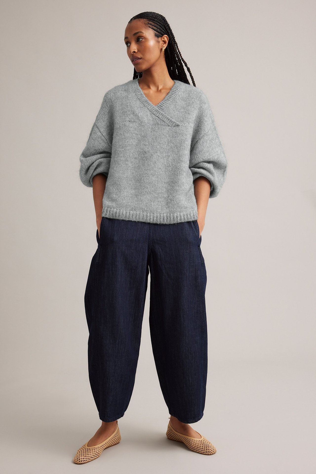 Framifrån, en kvinna i ljusgrå V-ringad stickad tröja, mörkblå vida byxor och beiga flätade skor.