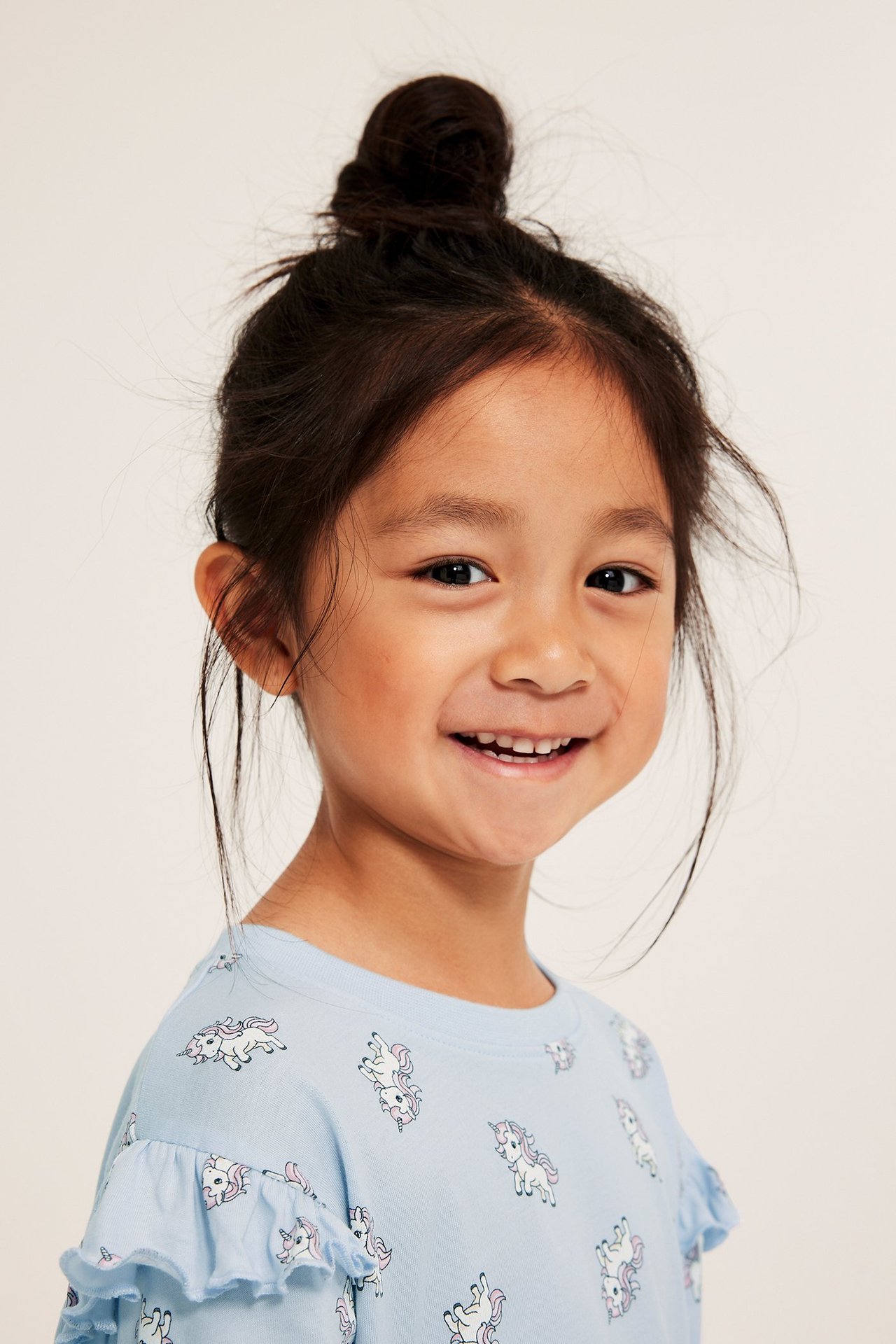 Lähikuva hymyilevästä lapsesta yllään vaaleansininen yksisarviskuvioinen röyhelöhihainen puuvillapyjama.