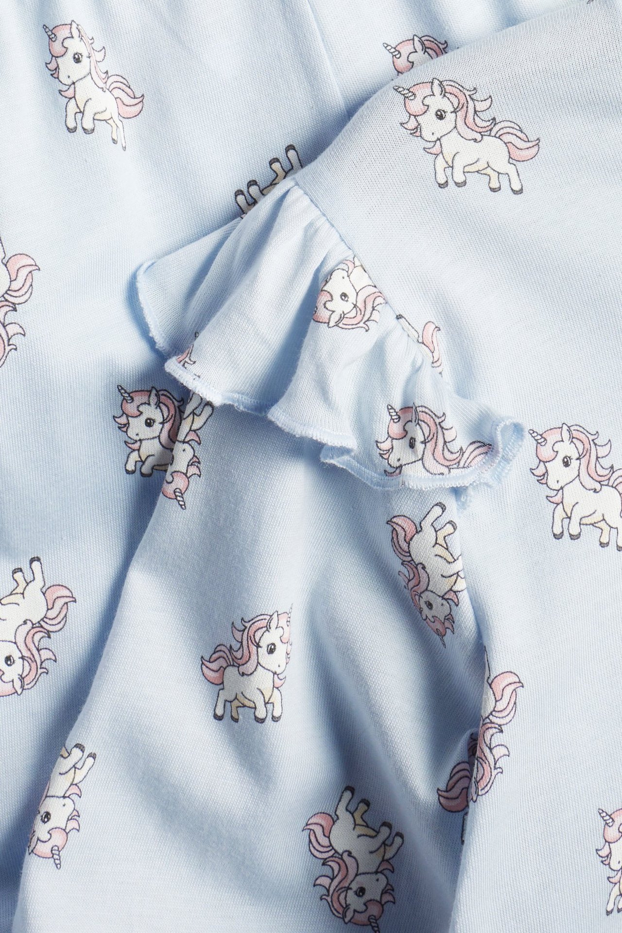 Zbliżenie na jasnoniebieską bawełnianą tkaninę piżamy z falbanką i wzorem w białe i różowe jednorożce.