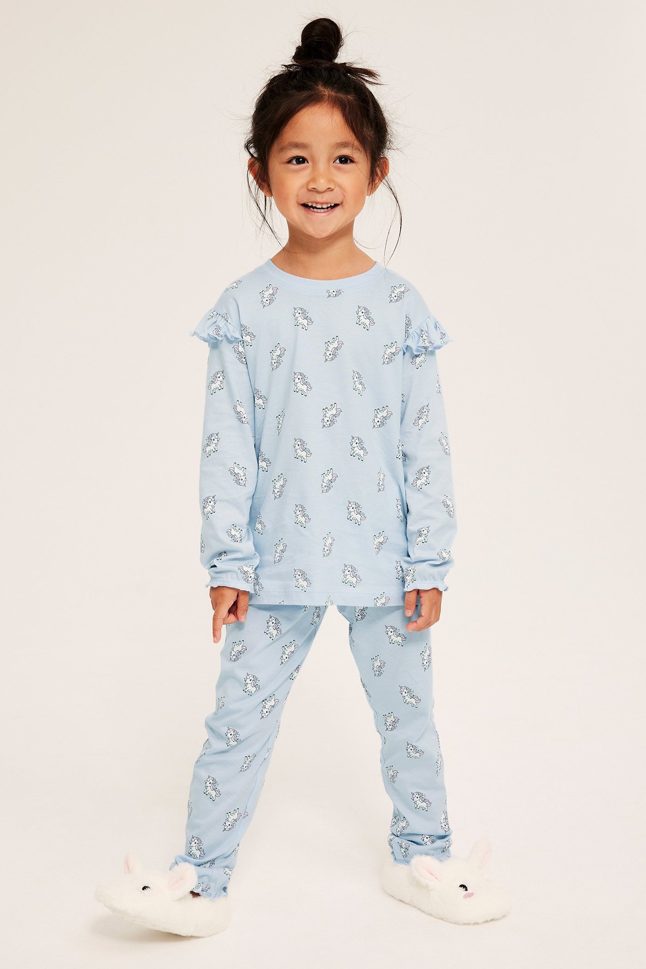 Widok z przodu dziecka w jasnoniebieskiej wzorzystej piżamie bawełnianej z falbanami i białymi kapciami w kształcie króliczków.