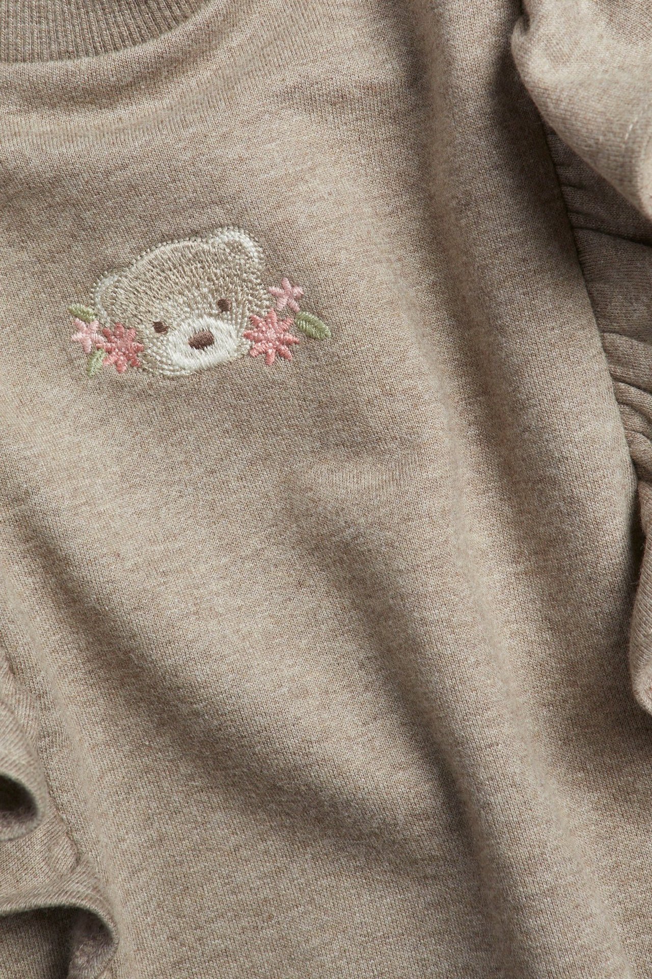 Nærbilde av en barnesweatshirt i beigemelert bomull med volanger og brodert teddybjørn med rosa blomster.
