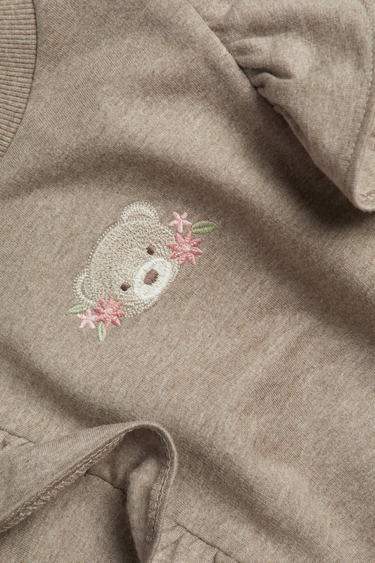 Zbliżenie brązowego melanżowego swetra dziecięcego z haftem misia i falbankami.