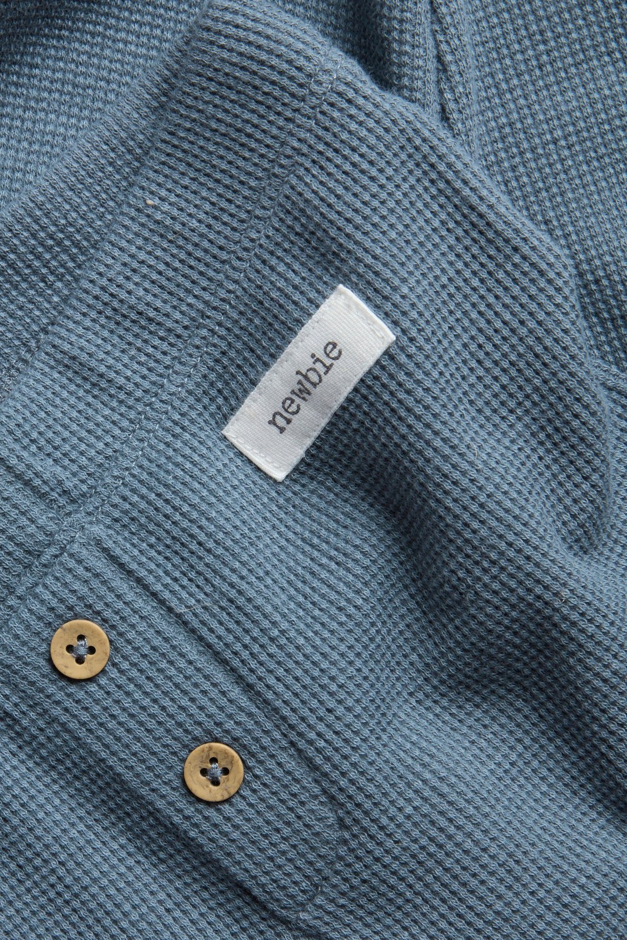 Zbliżenie niebieskich legginsów z dzianiny waflowej z ozdobnymi drewnianymi guzikami i białą metką Newbie.