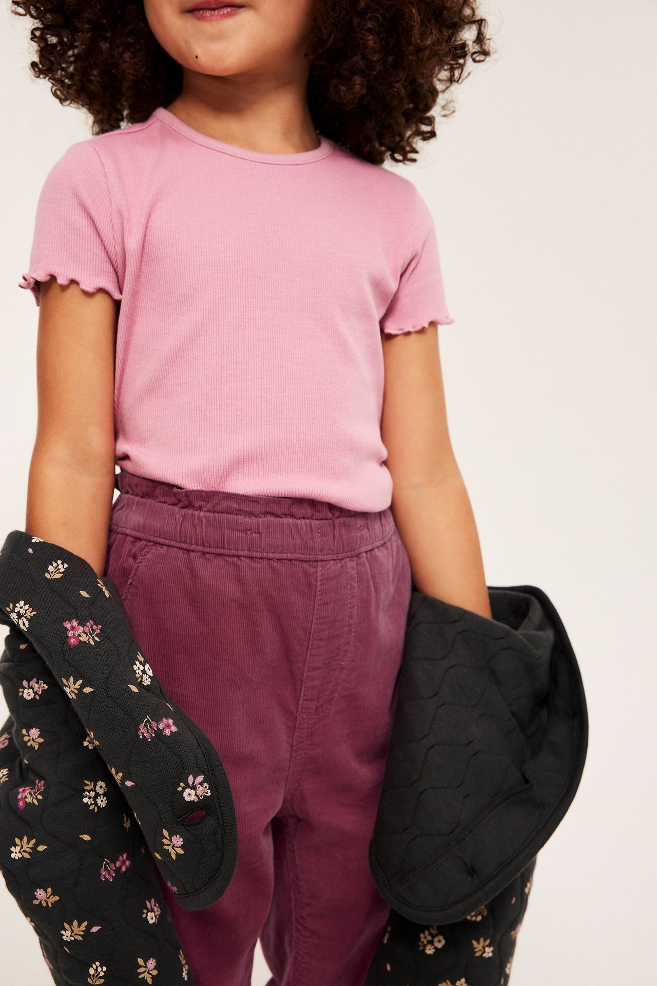 Frontbilde av et barn i mørkrosa kordfløyelbukser, en rosa t-skjorte, som holder svarte mønstrede grytekluter.