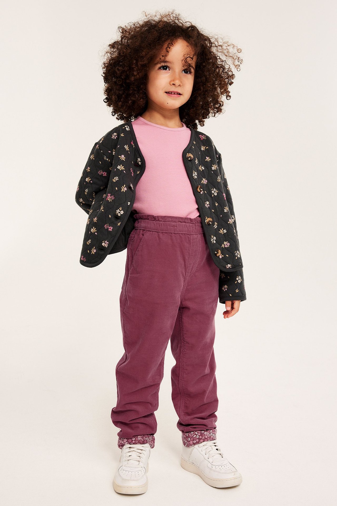 Frontalvisning: Barn i mørkrosa kordfløyelsbukser, rosa topp, svart blomstret cardigan og hvite joggesko.