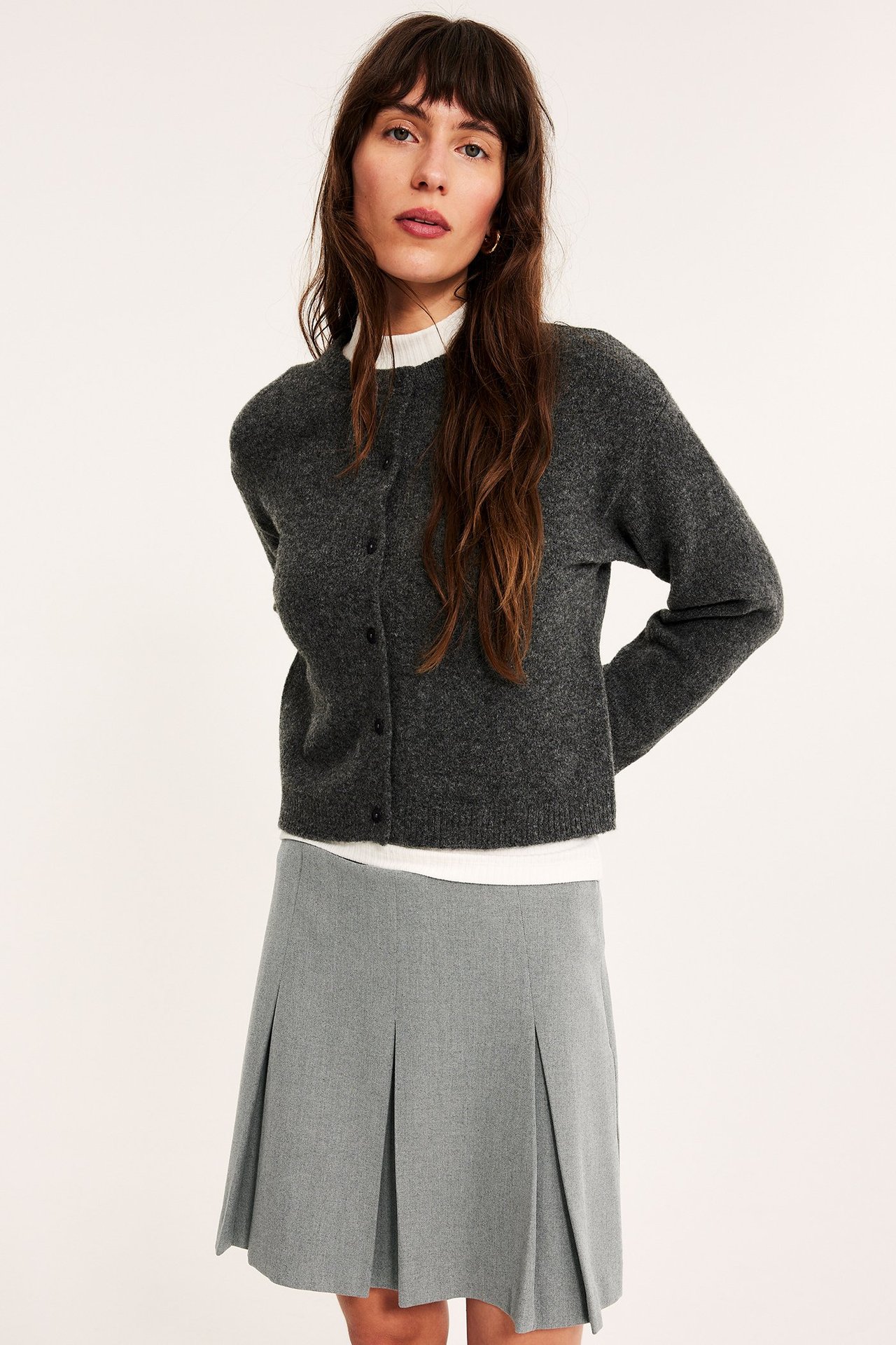 Forfra: Kvinne i lys grå melert plissert skjørt, mørk grå cardigan og hvit topp.