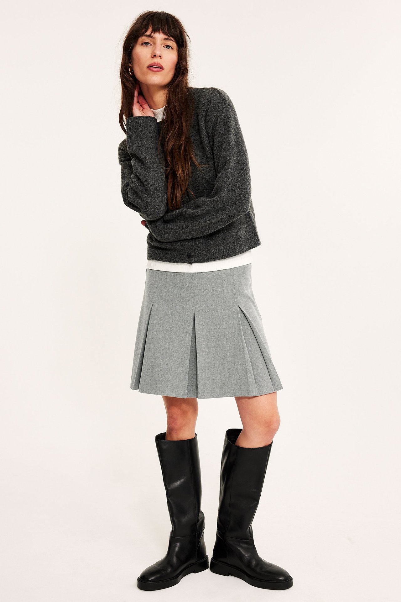Frontvisning: Kvinne i lys grå plissert melert skjørt, mørk grå cardigan, hvit topp og svarte knestøvler.