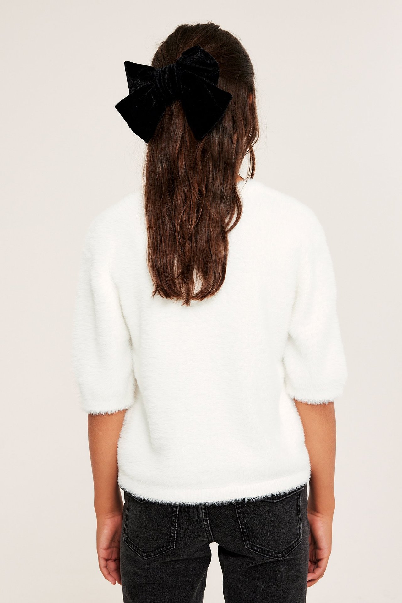 Ryggbilde av et barn i en offwhite fluffy genser, svarte jeans og en svart fløyelsløyfe i håret.