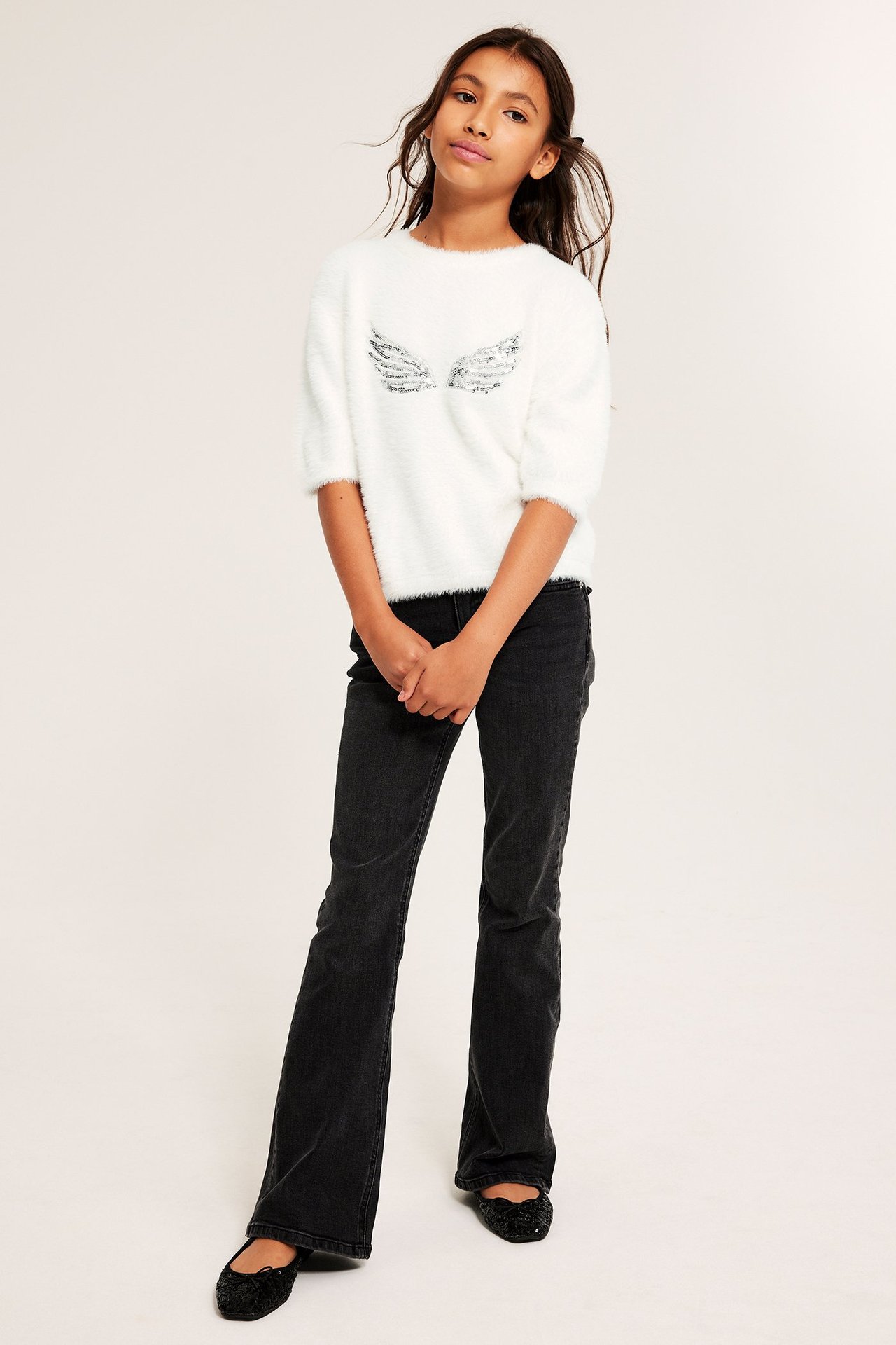 Frontvy av barn i offwhite fjädergarnströja med paljettvingar, svarta utsvängda jeans och ballerinaskor.