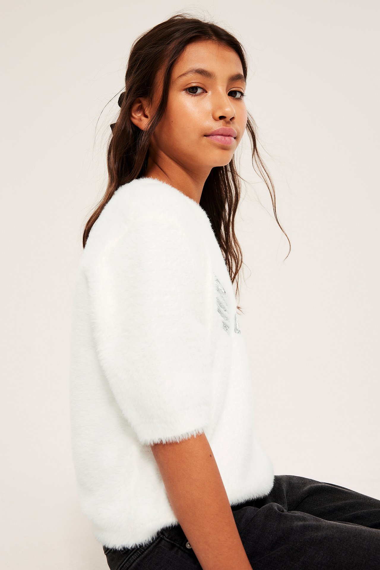 Sidebilde av et barn i offwhite fluffy strikket t-skjorte med korte ermer og paljettapplikasjon.