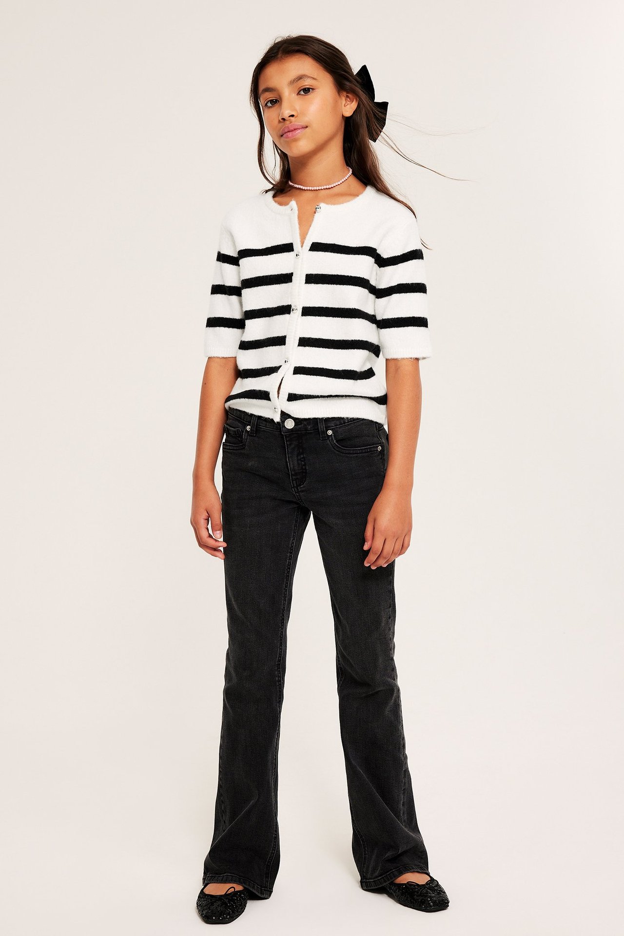 Frontvy: Barn i offwhite/svartrandig kortärmad knappkofta, svarta utsvängda jeans och svarta ballerinaskor.