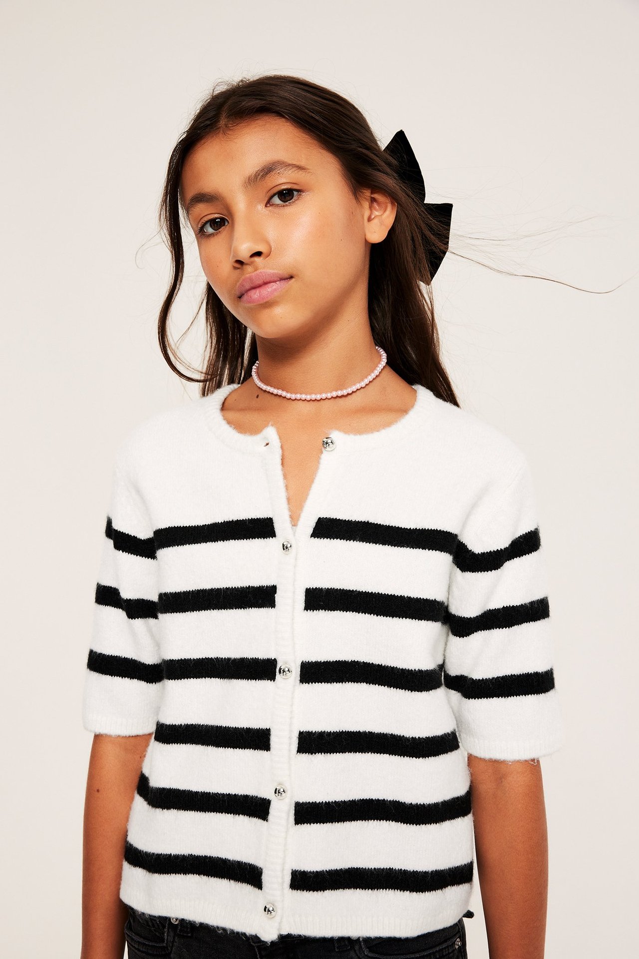 Barn bär offwhite-randig kortärmad kofta med dekorativa knappar, rosa halsband och svart hårrosett.